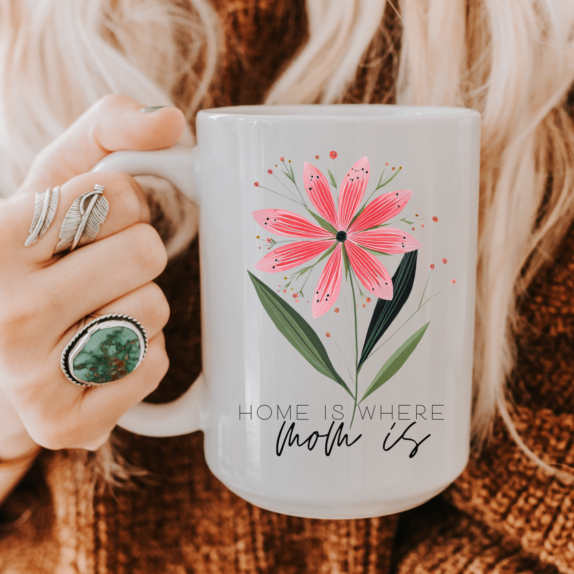 Person holding a 15oz white ceramic mug with a floral design and text 'Home is where mom is' on a textured surface.