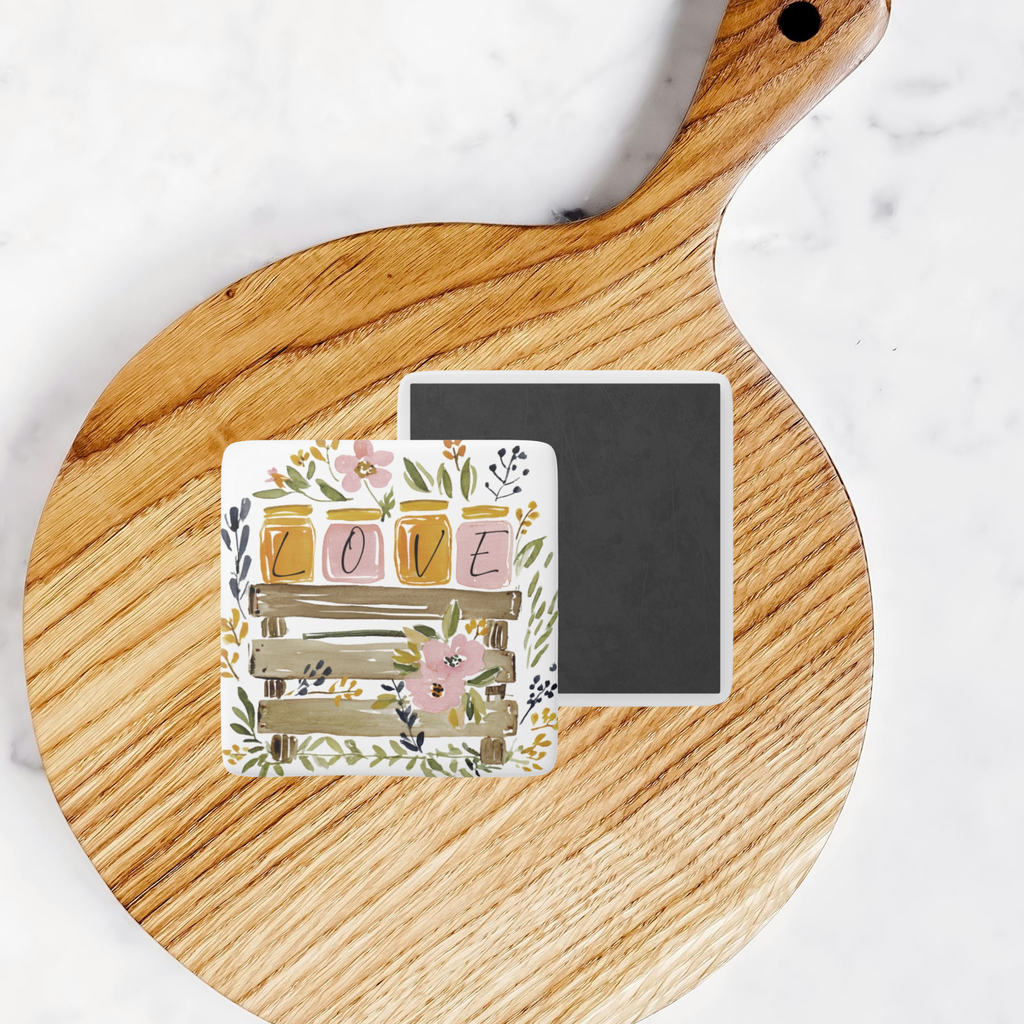 Front and back of decorative porcelain magnet with floral design on a wooden cutting board for size comparison.