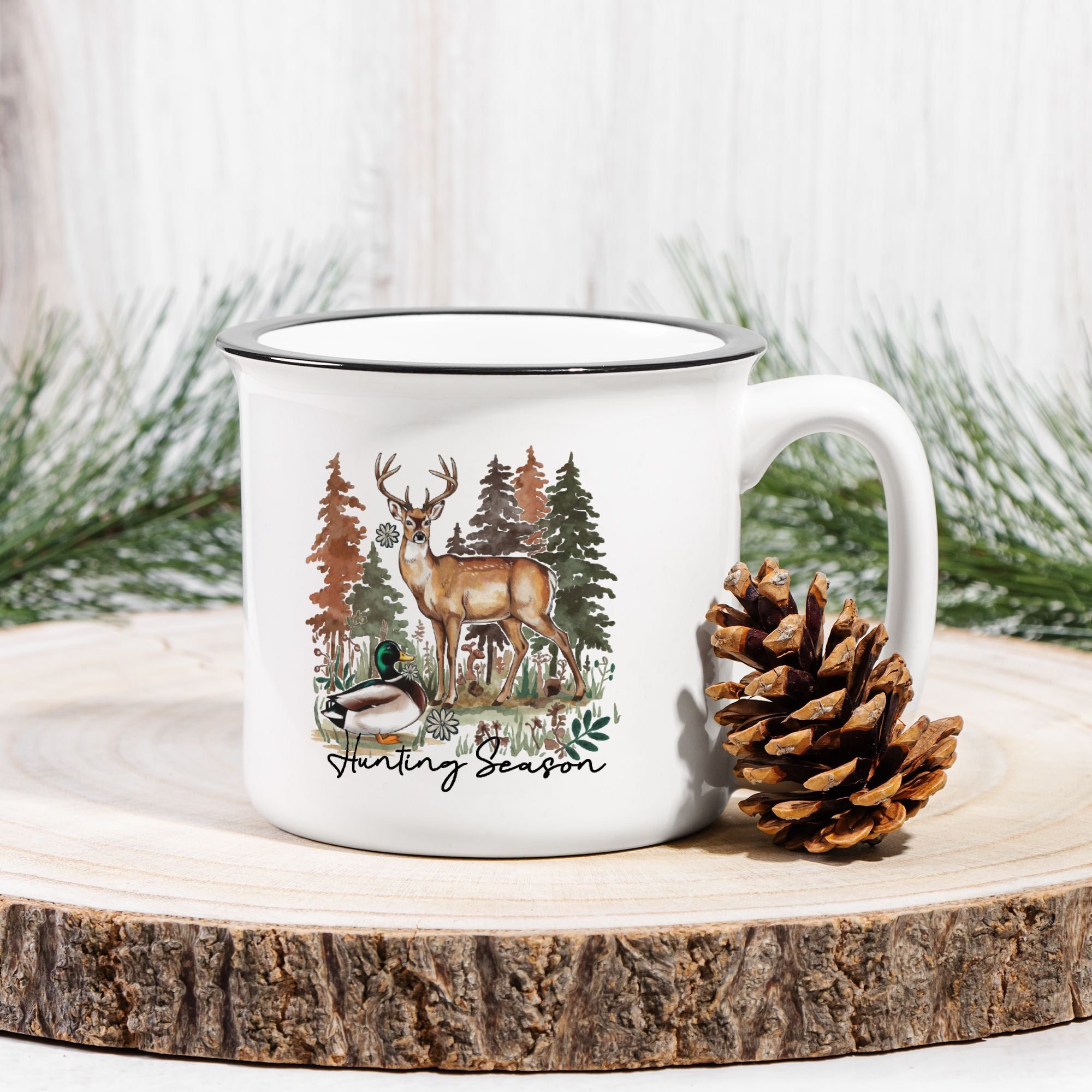 White ceramic camp mug with nature-themed design featuring a deer and trees, placed on a wooden surface with a pine cone and evergreen branches.