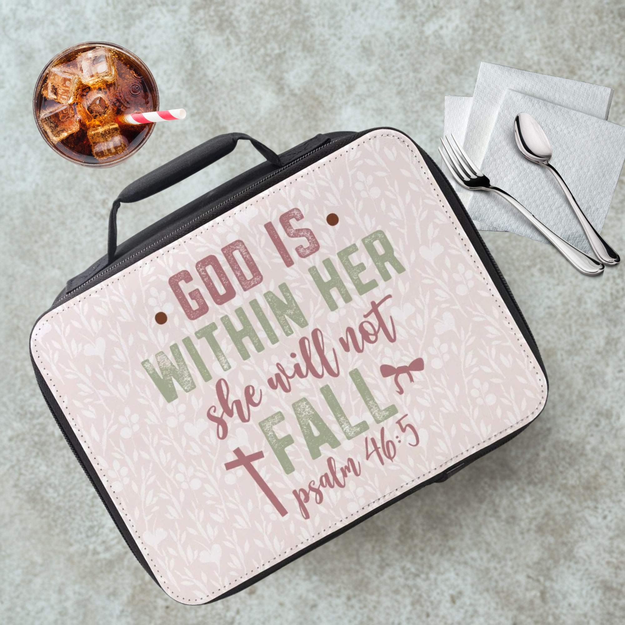 Lunch box with 'God is within her, she will not fall' quote on a concrete surface with a drink and utensils.
