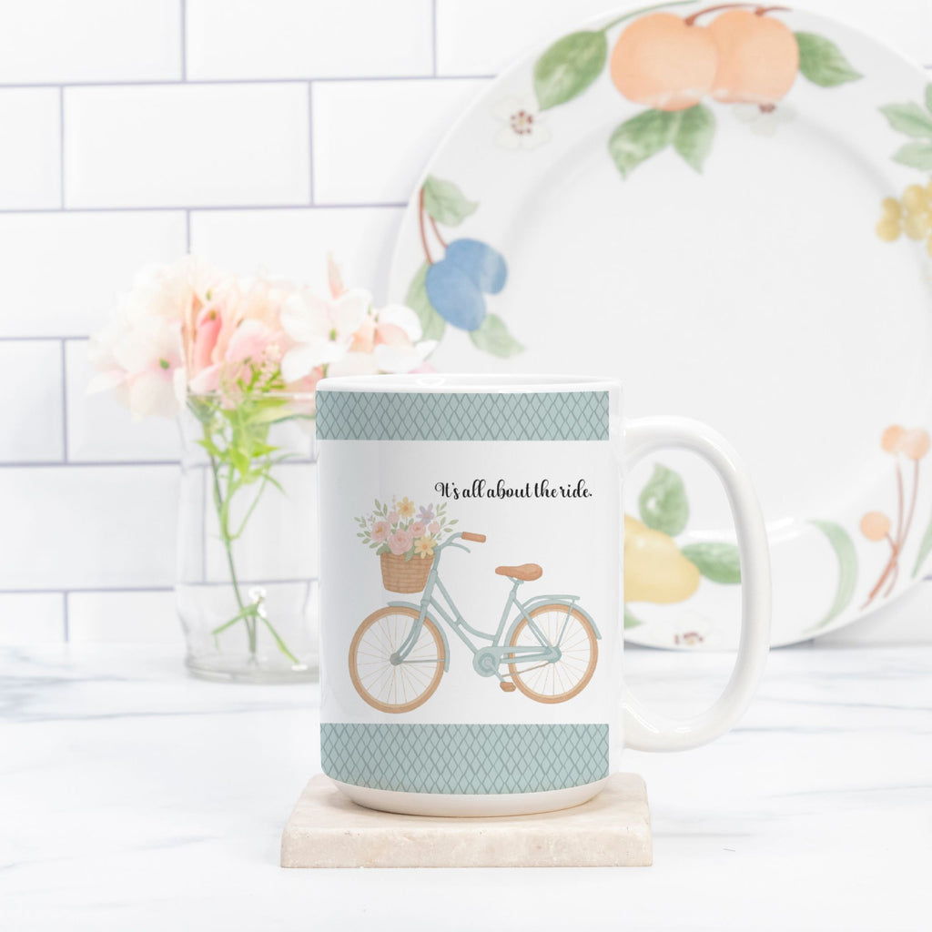 Mug with bicycle design and text 'It's all about the ride' on a white surface with a blurred background.