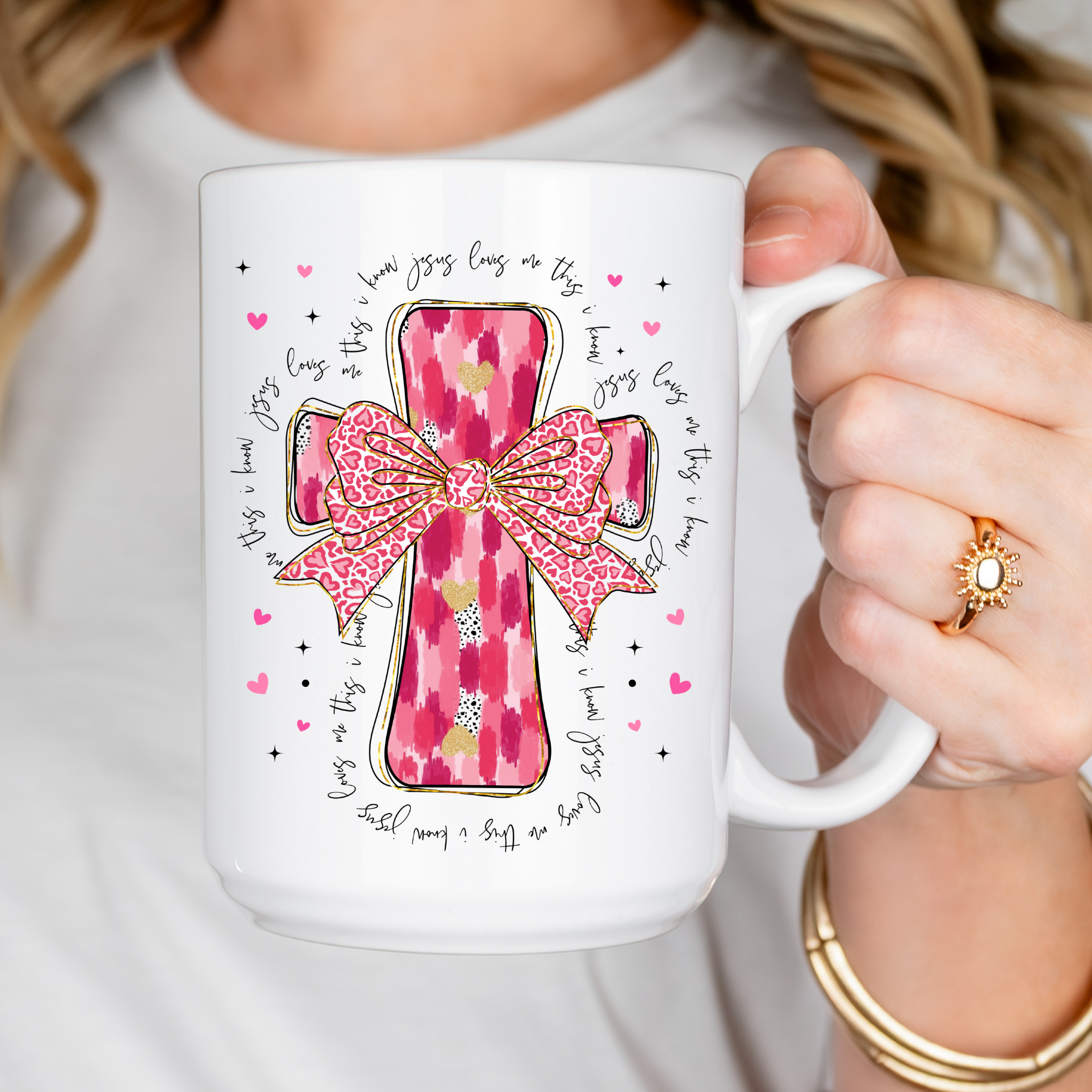 Person holding a mug with a pink cross and bow design with text 'Jesus Loves Me'.