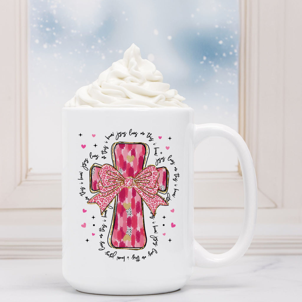 White mug with pink cross design and text, filled with hot chocolate and whipped cream.