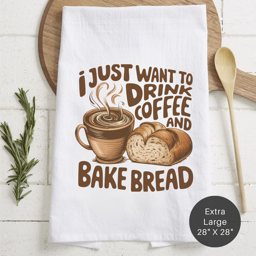 White tea towel with coffee and bread design and text ' I Just Want To Drink Coffee And Bake Bread' on a wooden surface.