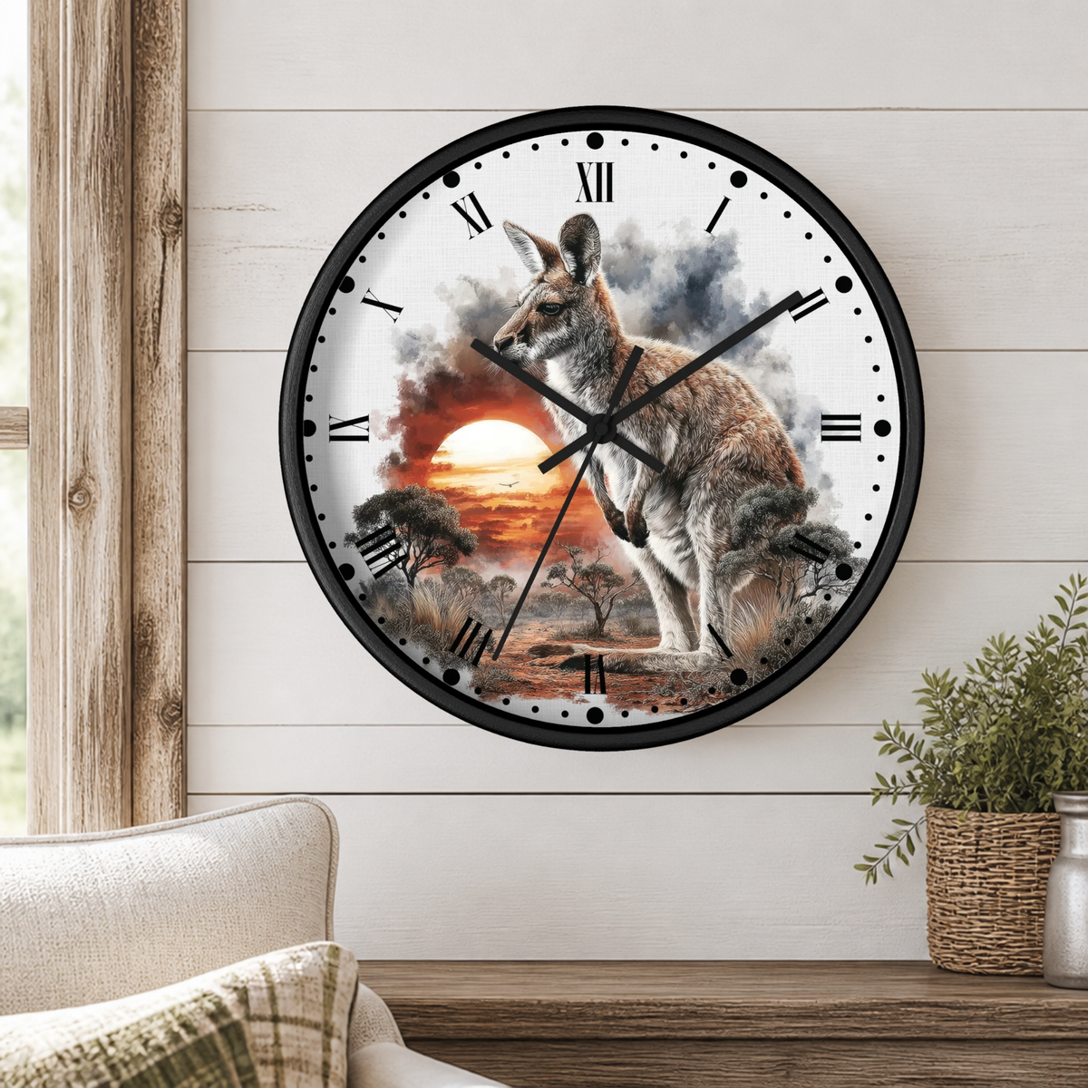 A round wall clock with a black frame and a printed kangaroo landscape design, mounted on a white wood-paneled wall above a wooden shelf with a plant and part of a sofa visible.