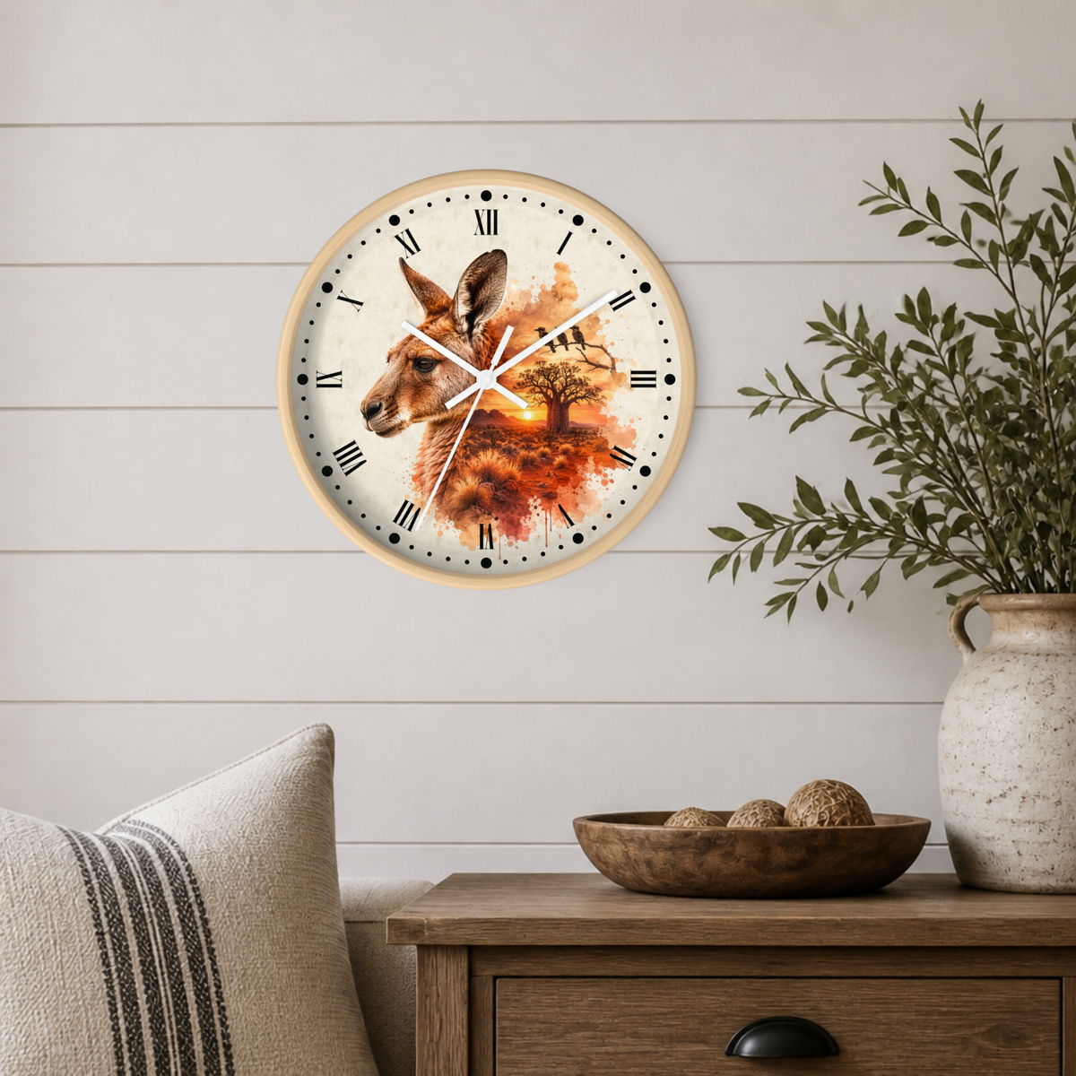A round wall clock with a light wood frame hangs on a white shiplap wall above a wooden side table. The clock face features a watercolor-style illustration of a kangaroo in warm autumn tones with a tree and sunset.