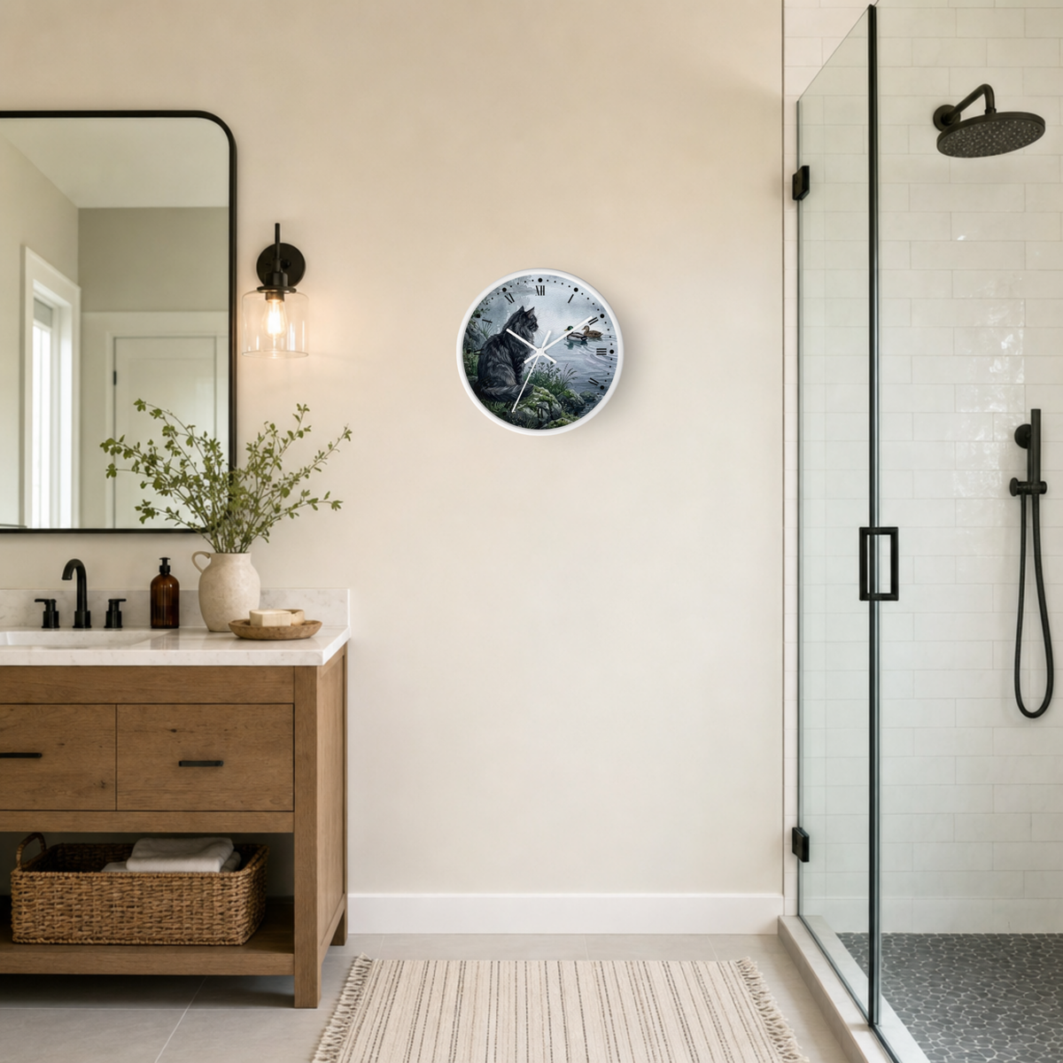 A round wall clock with a cat on the bank of a lake watching two ducks, print hangs on a beige wall in a modern bathroom next to a wooden vanity and glass shower enclosure.