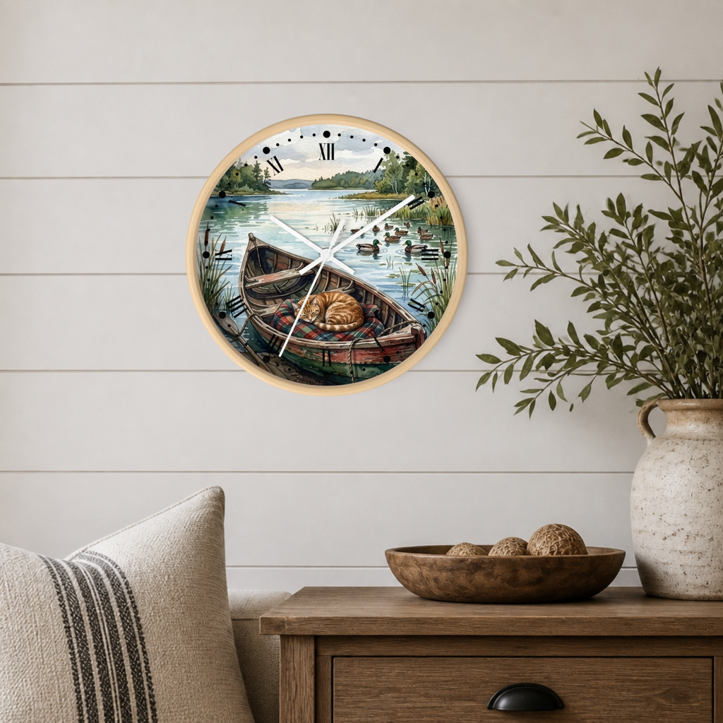 Round wall clock with a light wood frame and a scenic illustration of a cat sleeping in a rowboat on the face, mounted on a white shiplap wall above a wooden side table with a vase and bowl.