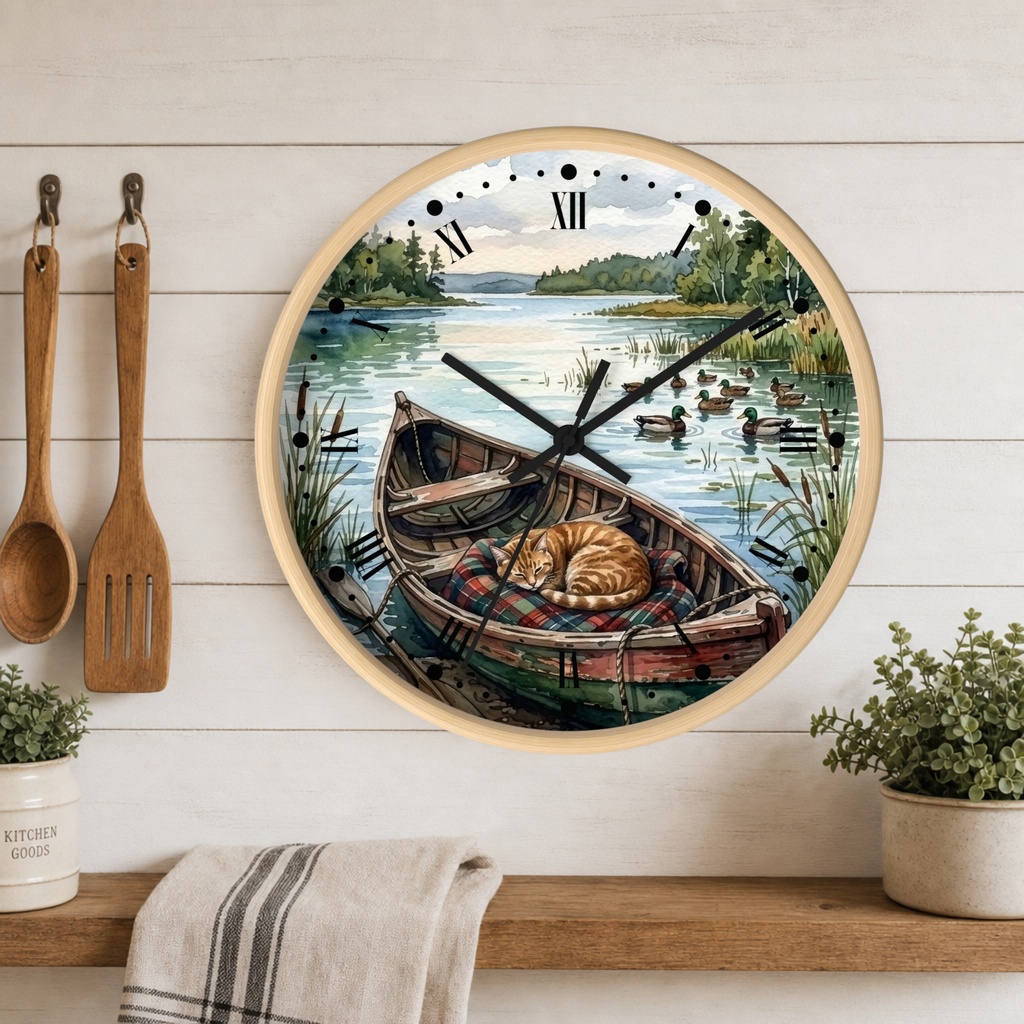 Round wall clock with a light wood frame featuring a scenic illustration of a cat sleeping in a rowboat on a lake, displayed on a white wood plank wall above a shelf with kitchen utensils and plants.