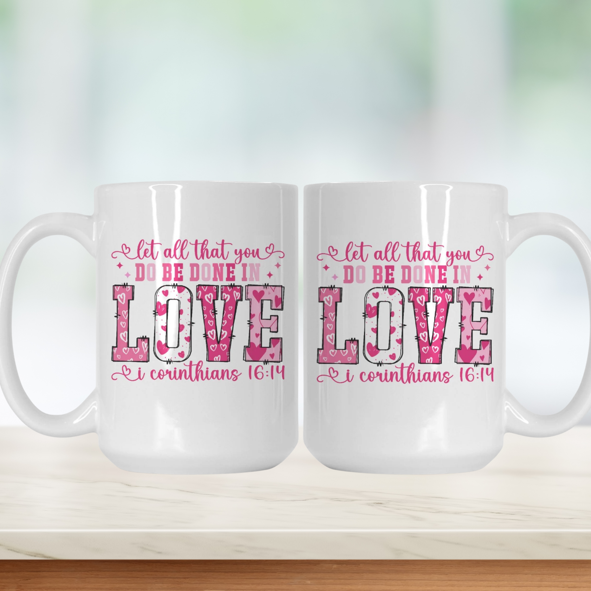 Two white mugs with pink 'let all that you do be done in love' text on a light wooden surface.