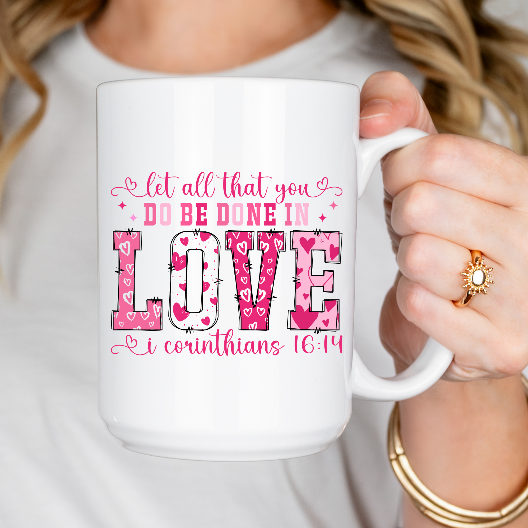 White mug with pink and red text held by a person wearing a gold ring and bracelet.