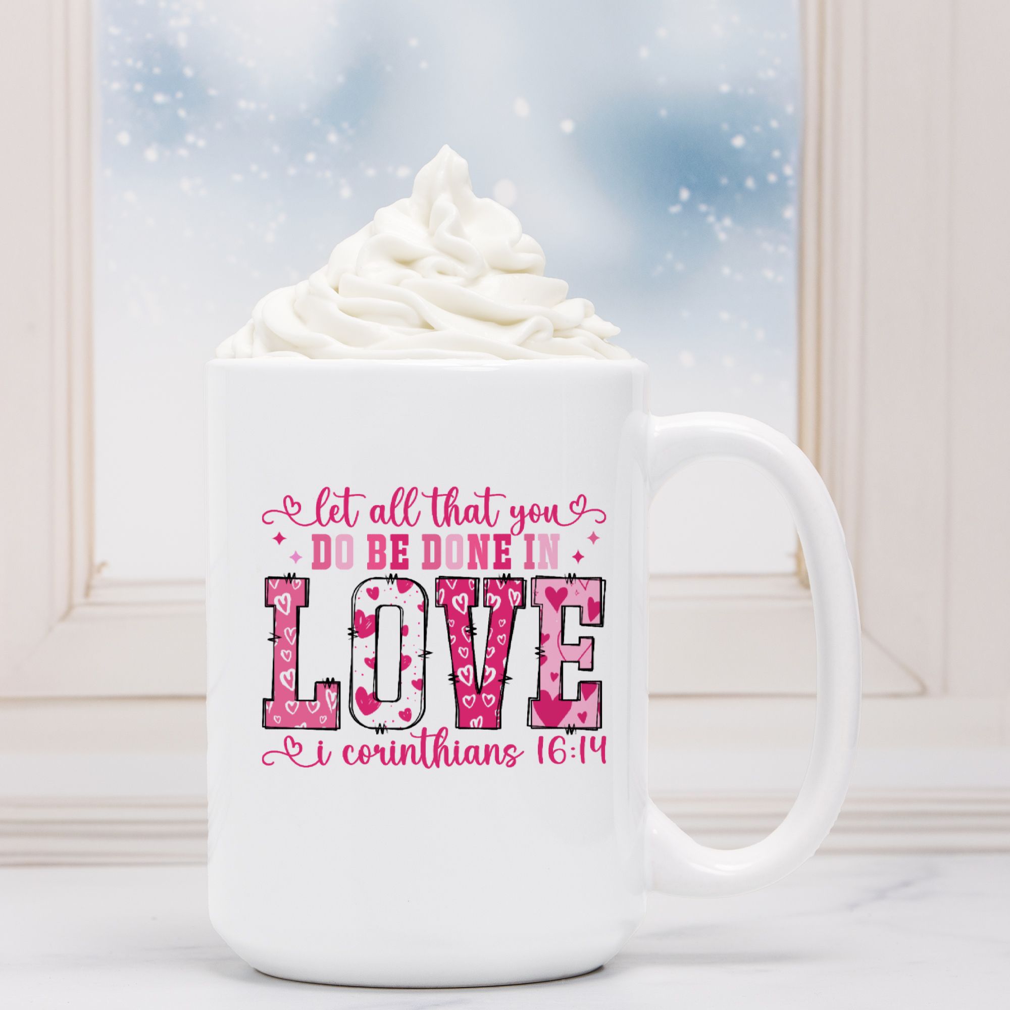 White mug with pink text and design, filled with hot chocolate and whipped cream, against a snowy window background.