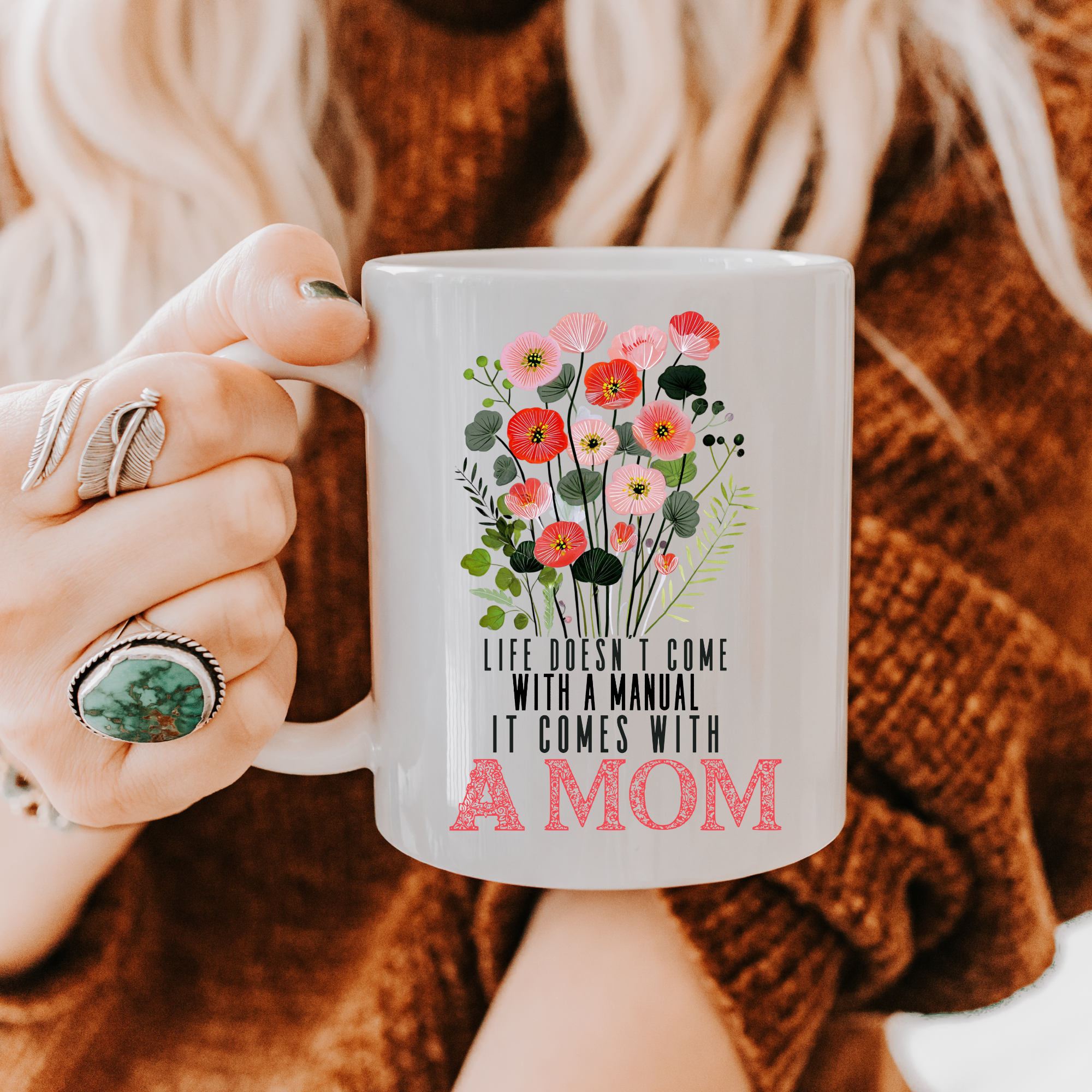 Person holding an 11oz white mug with floral design and text 'Life doesn't come with a manual it comes with A MOM', wearing a brown sweater.