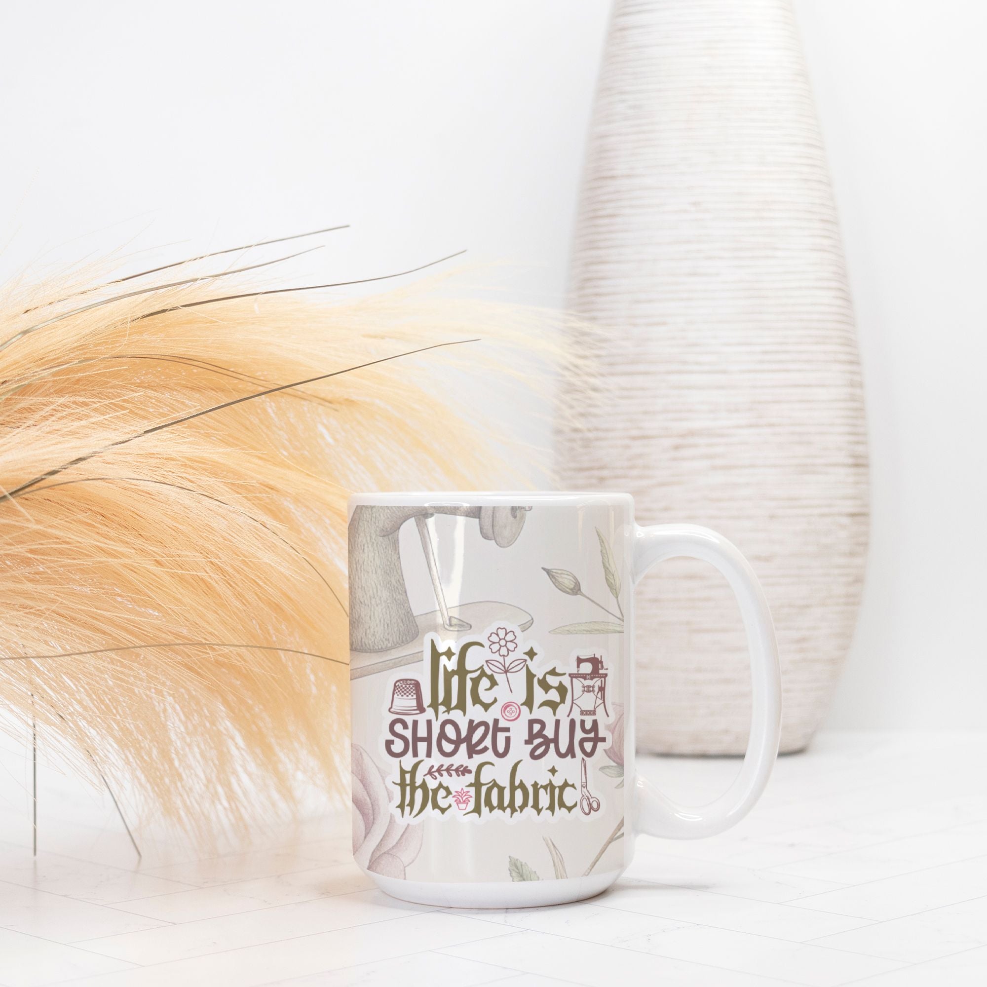 Mug with 'Life is short, buy the fabric' text on a white surface with decorative elements.