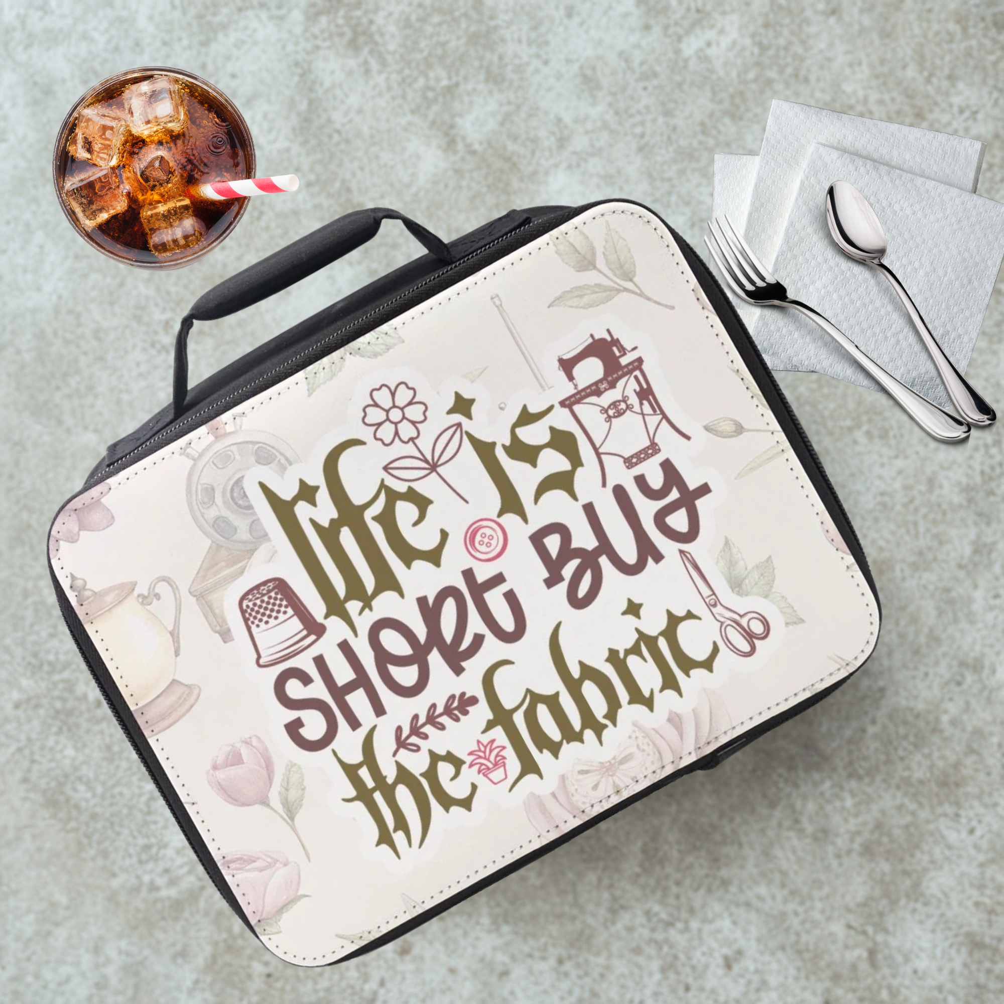 Lunch bag with 'Life is short, buy the fabric' text on a cafeteria table with a drink and utensils.