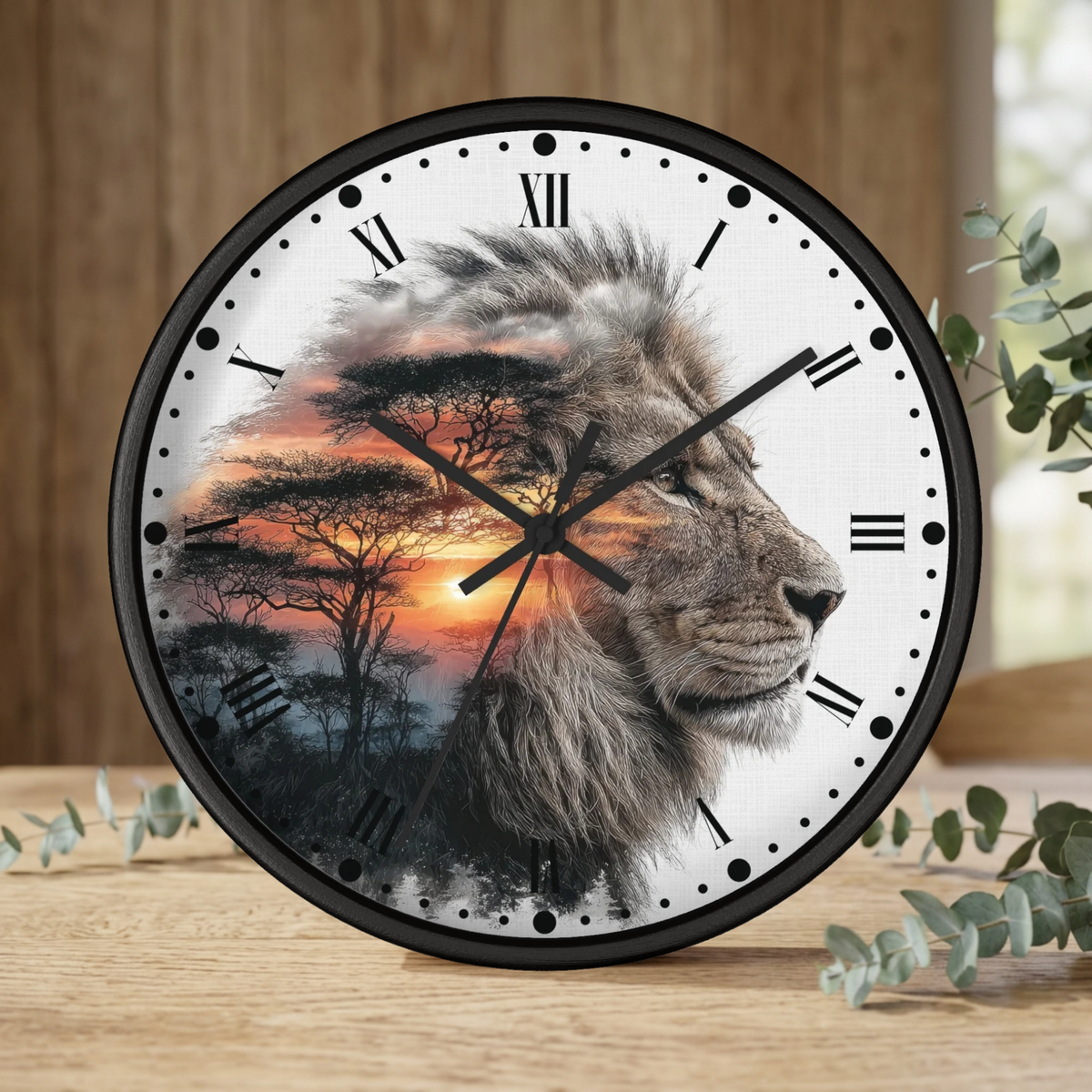 A round wall clock with a black frame and a white face featuring a lion profile over an African savanna sunset scene with trees. The clock is shown on a wooden surface with eucalyptus leaves and a blurred wood-paneled background.