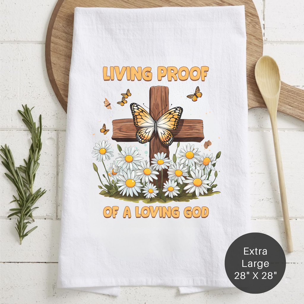 White tea towel with a cross, butterfly, and flowers design and text 'Living Proof Of A Loving God' on a wooden surface.
