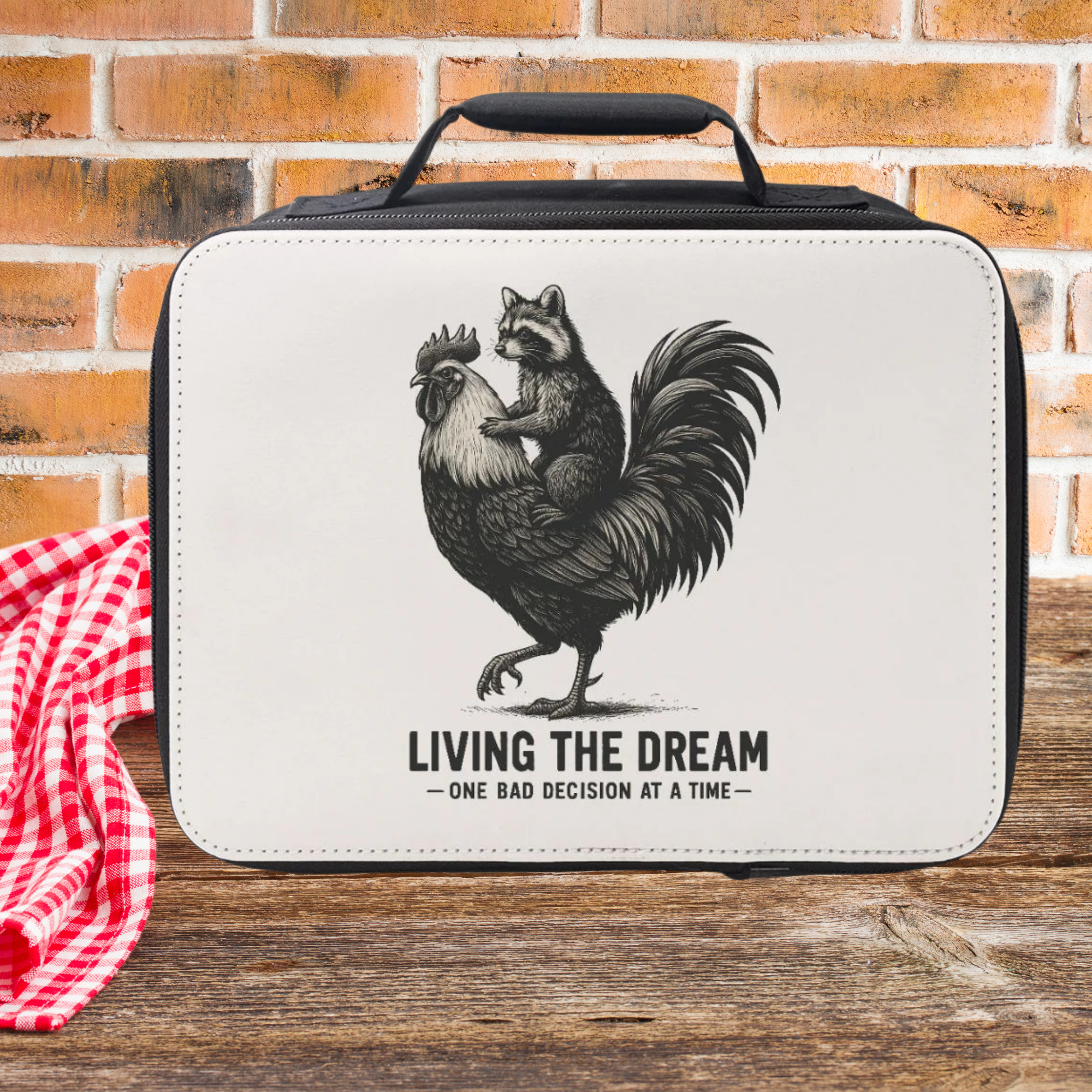 Lunch bag with a graphic of a raccoon on a rooster and text 'Living the Dream' on a wooden picnic table against a brick wall background.
