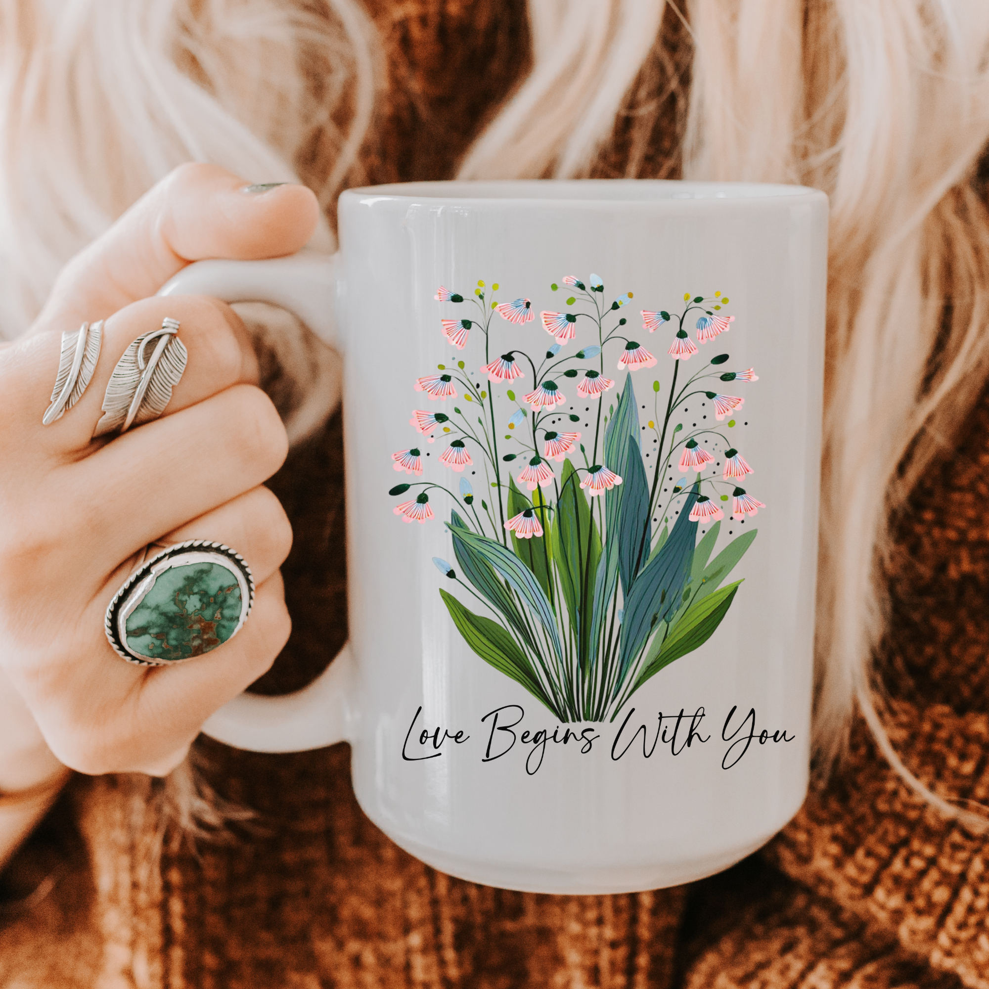 Person holding a 15oz. mug with floral design and text 'Love Begins With You' on a textured surface.