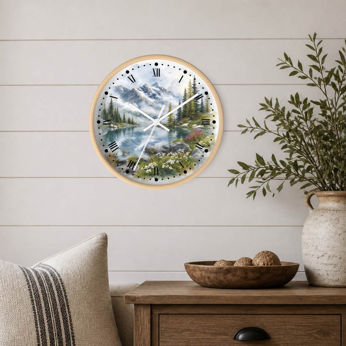 A round wall clock with a light wood frame and a scenic mountain lake print, mounted on a white shiplap wall above a wooden side table with a vase and bowl.