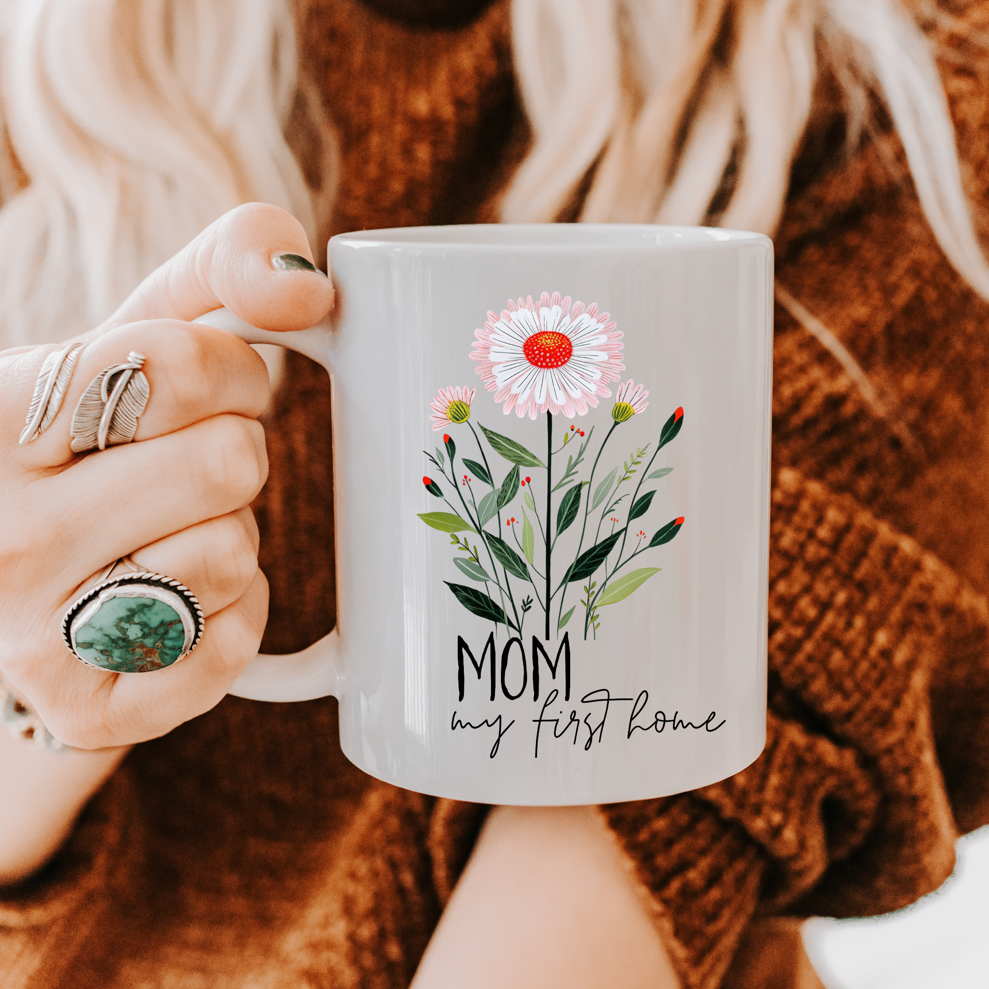 Person holding an 11oz mug with floral design and 'Mom my first home' text, wearing a brown sweater.