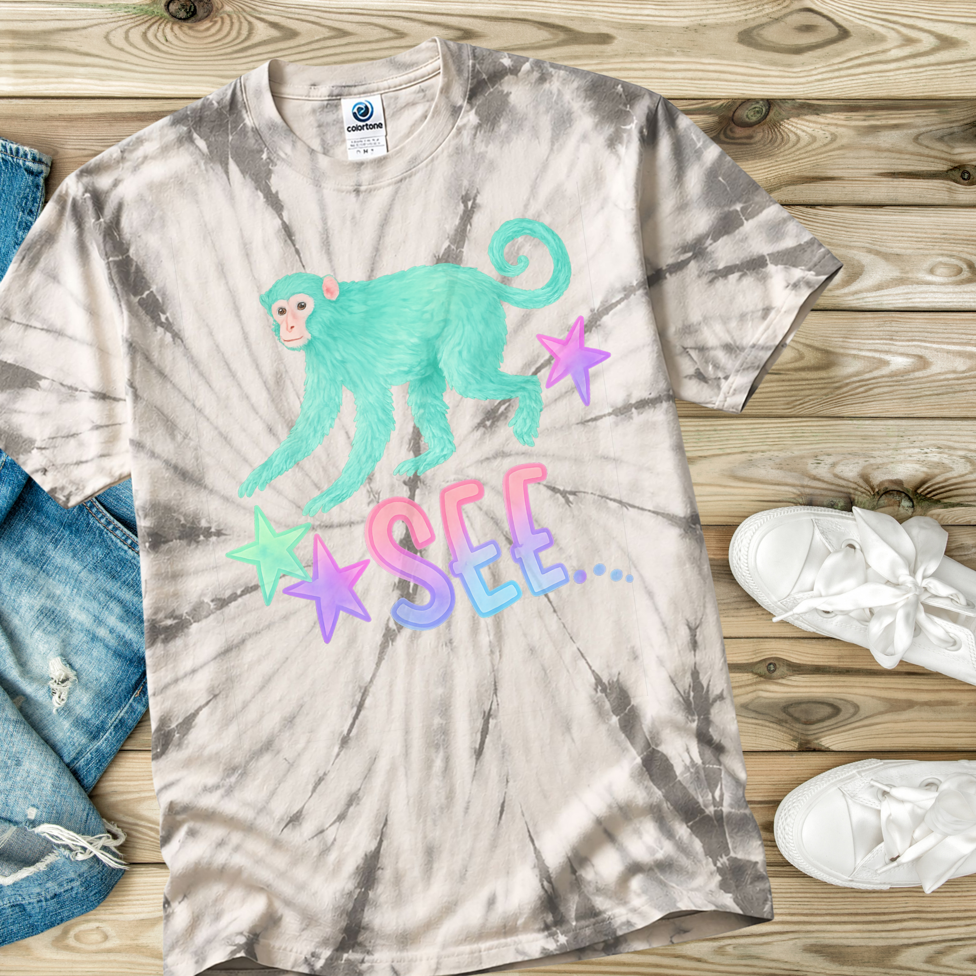 Tie-dye t-shirt with a monkey graphic and 'SEE' text on a wooden surface with jeans and shoes.