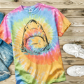 Tie-dye t-shirt in the color Eternity with a hand drawn shark design on a wooden surface with jeans and shoes.
