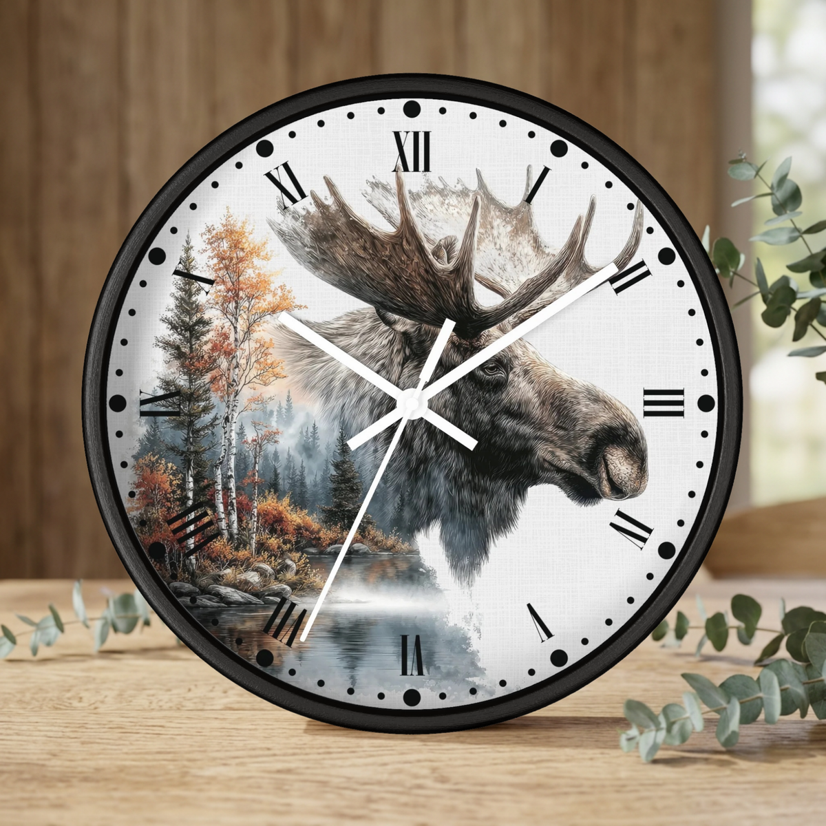 A round wall clock with a black frame and a white face featuring a moose head over a forest landscape, styled on a wooden surface with eucalyptus leaves.
