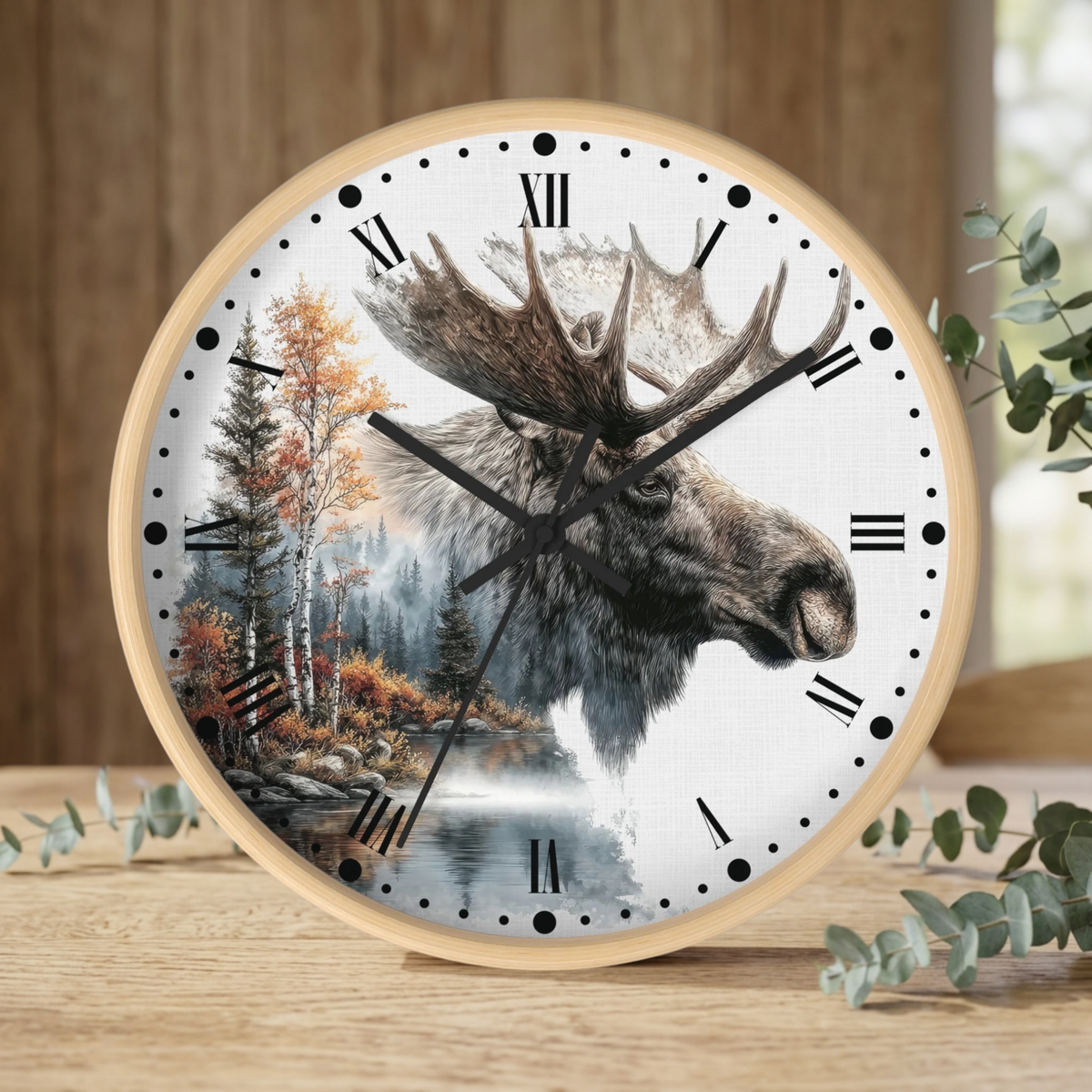 A round wall clock with a light wood frame and a white face featuring a moose head over a forest landscape, styled on a wooden surface with eucalyptus leaves.