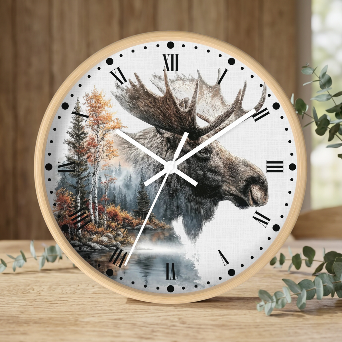 A round wall clock with a light wood frame and a white face featuring a moose head over a forest landscape, styled on a wooden surface with eucalyptus leaves.