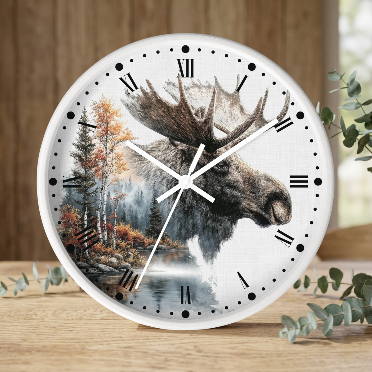Round wall clock with a white frame and a printed moose head over a forest landscape scene, with black Roman numerals and white hands, shown on a wooden surface with blurred greenery in the background.