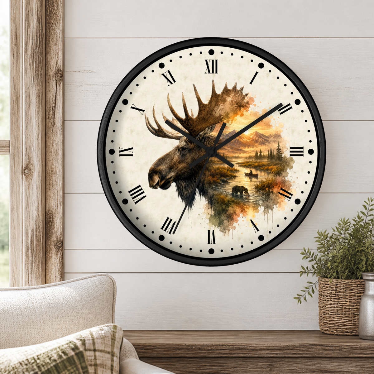 A round wall clock with a black frame and Roman numerals, featuring a moose head and autumn landscape artwork on the face, mounted on a white shiplap wall above a wooden mantel with a plant.
