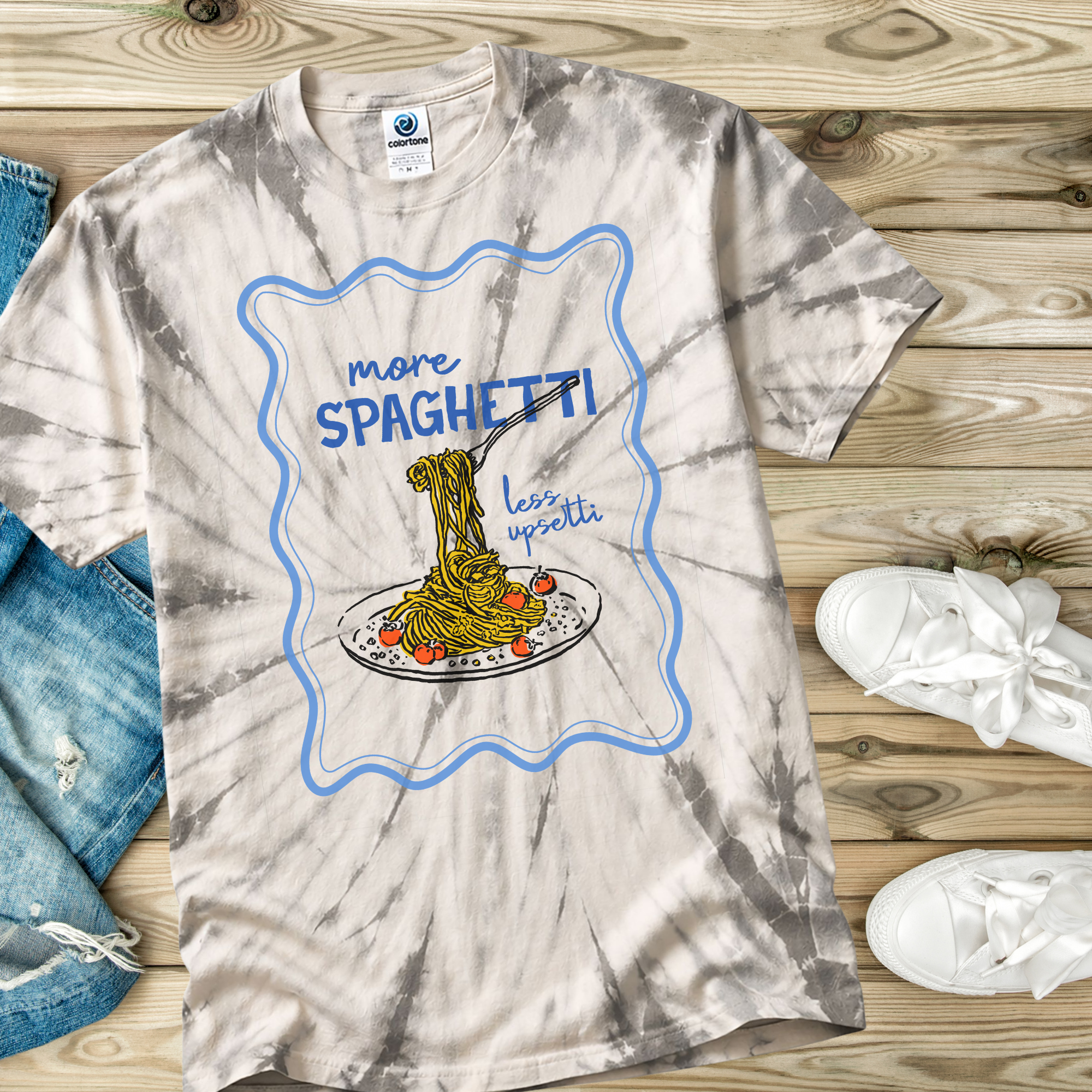 Tie-dye t-shirt with spaghetti graphic and text on a wooden surface with jeans and shoes.