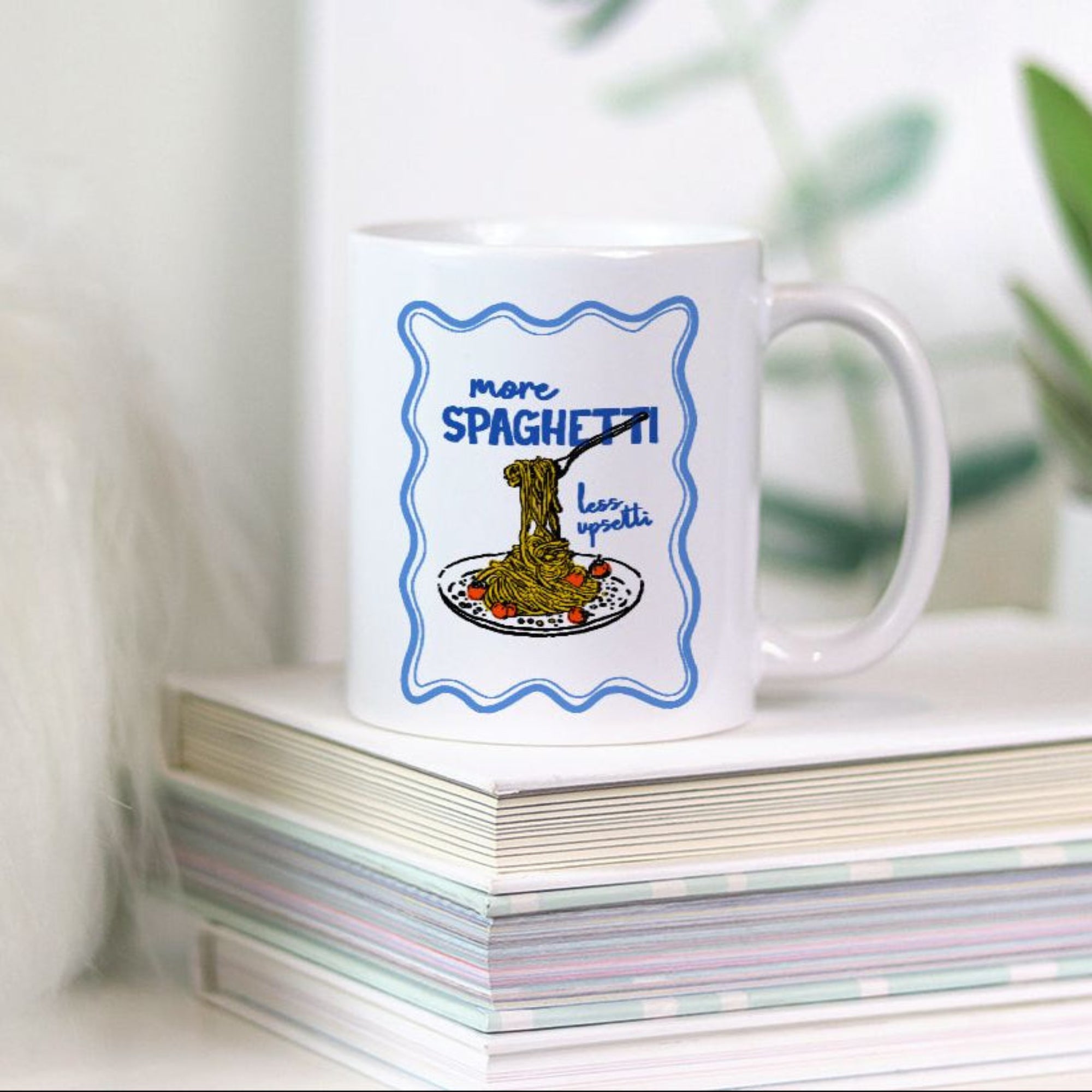 White 11oz. mug with spaghetti design on a stack of books with a plant in the background
