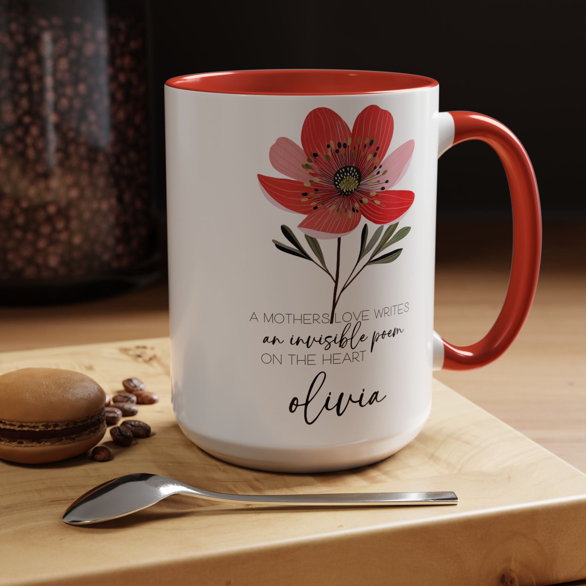 White mug with red interior and handle, featuring a floral design and text, on a wooden surface with coffee beans and a spoon.