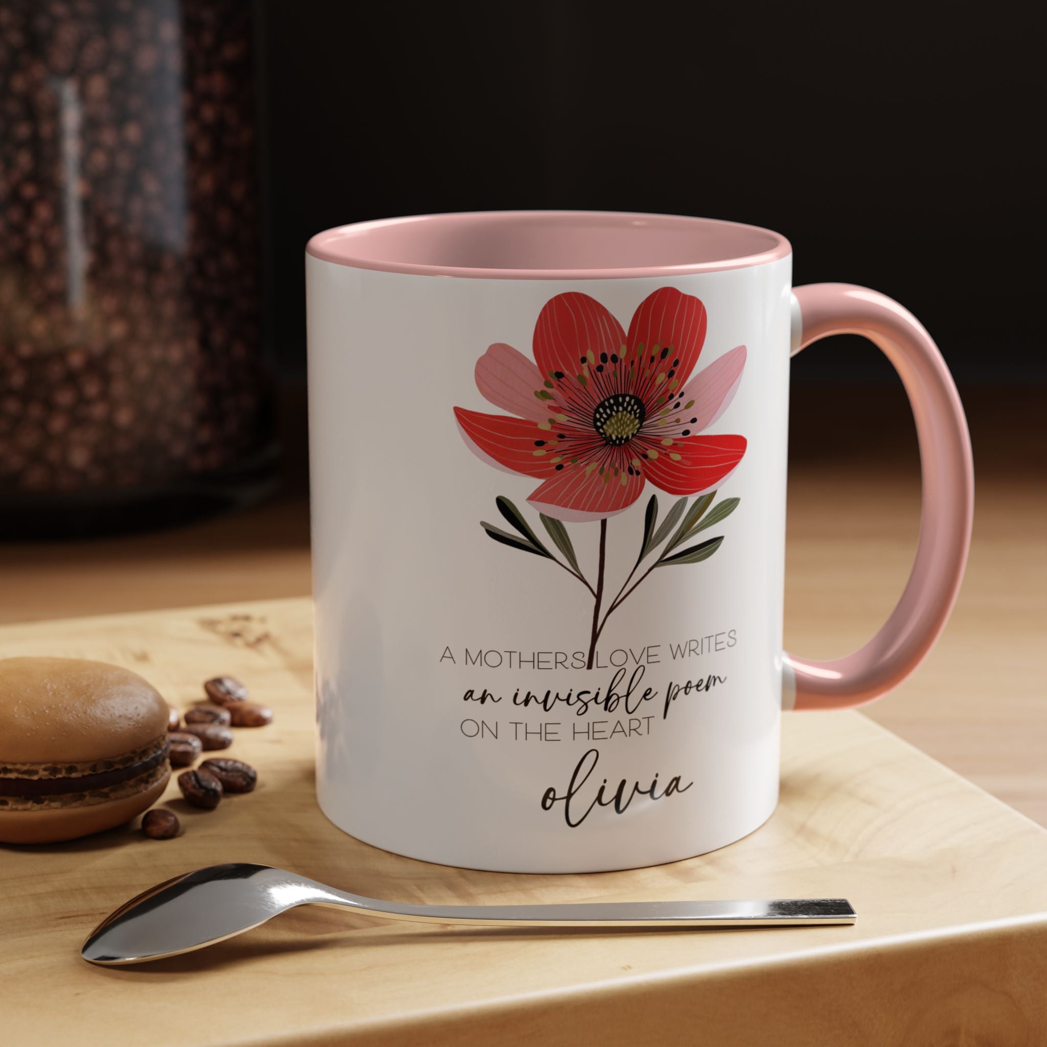 White mug with pink interior and handle, featuring a red flower design and text, on a wooden surface with coffee beans and a spoon.