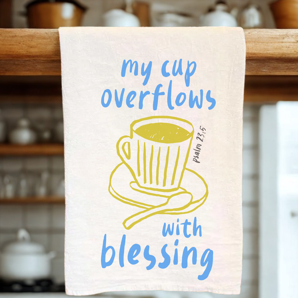 Kitchen towel with cup and saucer design and text 'my cup overflows with blessing' on a wooden rack.