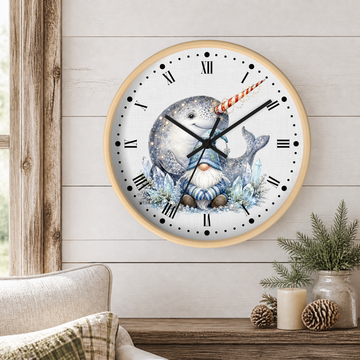 A round wall clock with a light wood frame hangs on a white shiplap wall above a wooden table. The clock face features a whimsical illustration of a narwhal and a small gnome, with black Roman numerals and black clock hands.