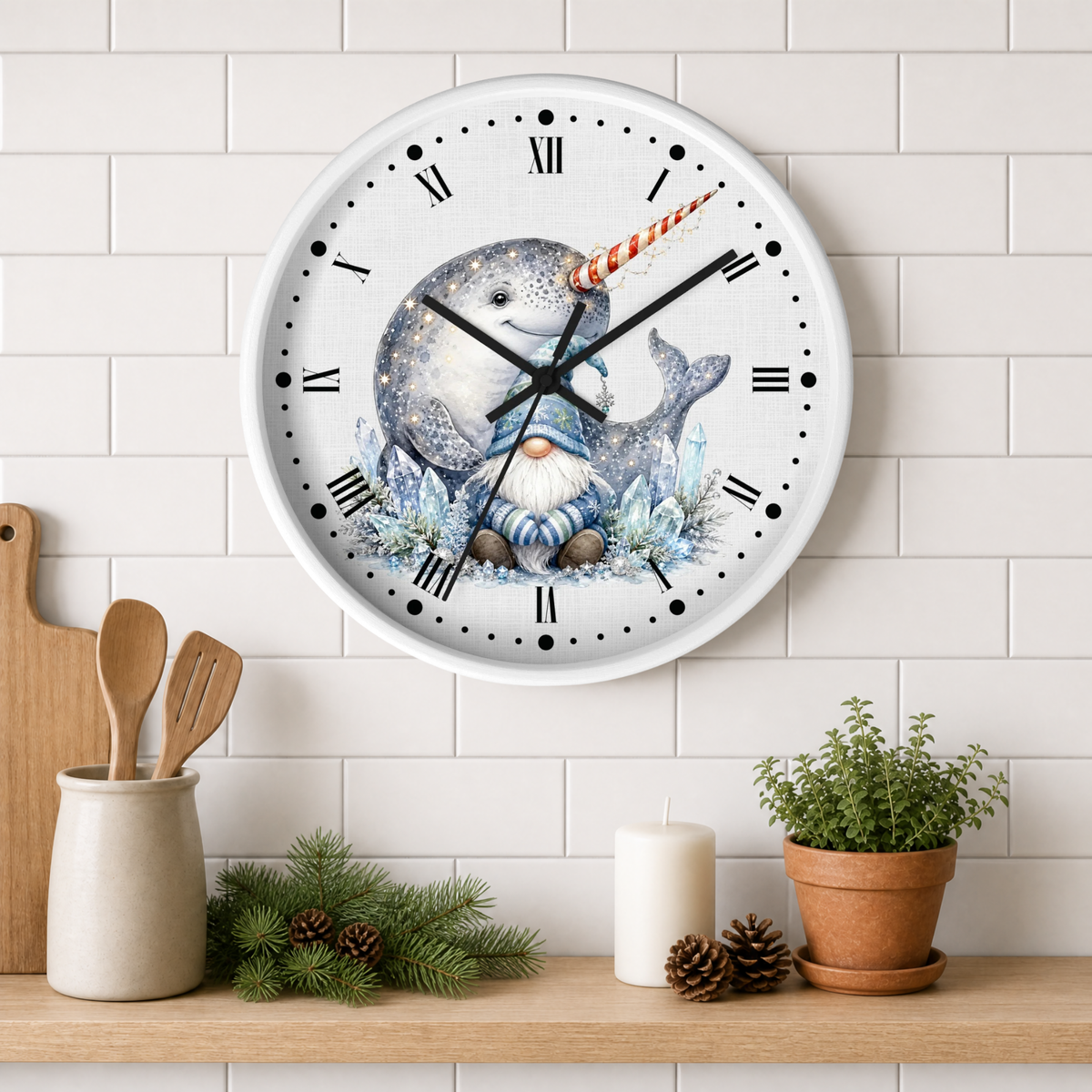 A round wall clock with a white face and Roman numerals, featuring an illustration of a blue-gray narwhal with a striped horn and a small gnome at the bottom. The clock is mounted on a white subway tile wall above a wooden shelf styled with kitchen utensils, pine branches, a candle, and a potted plant.