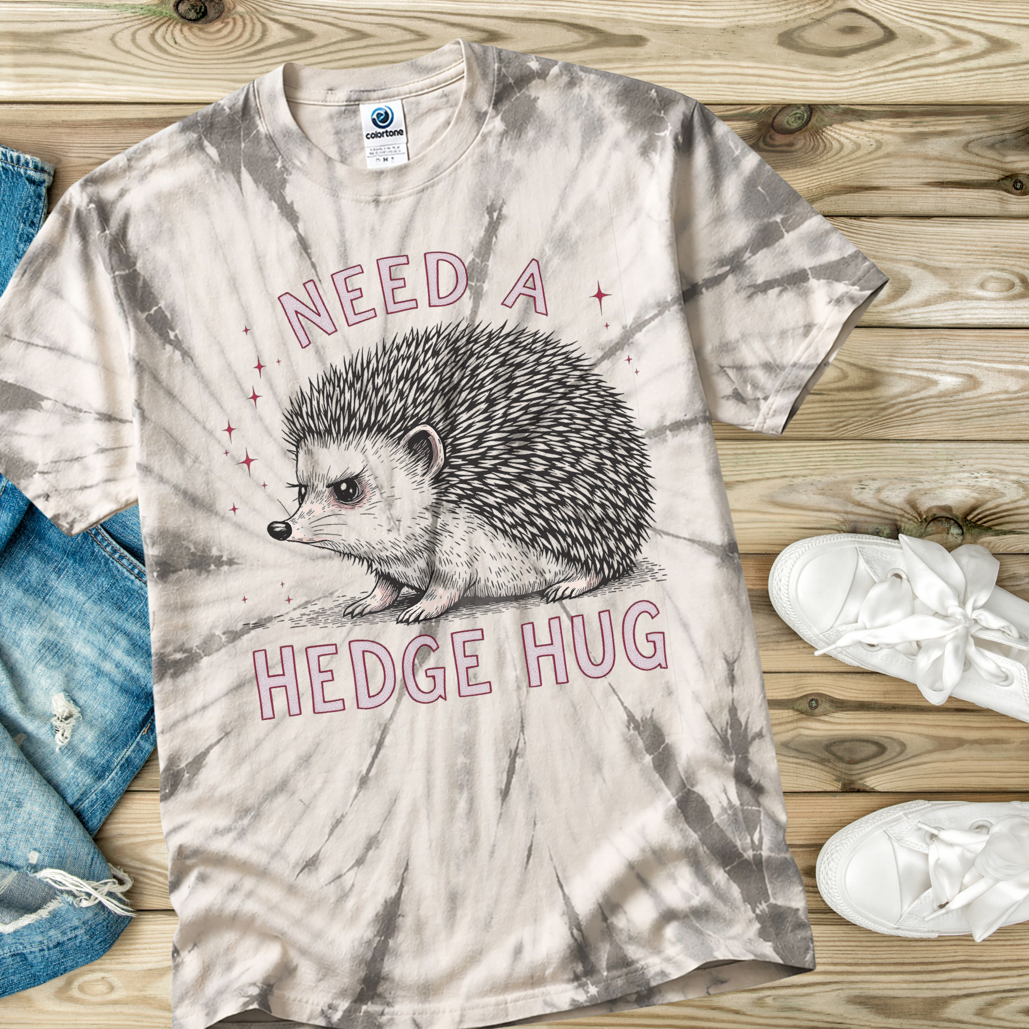 Tie-dye t-shirt with hedgehog graphic and 'Need a Hedge Hug' text on a wooden surface.