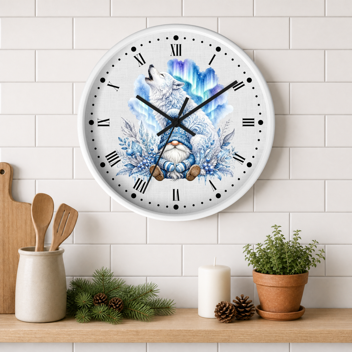 A round wall clock with a white face and black Roman numerals, featuring a winter illustration of a white wolf howling at a glowing blue aurora with a small gnome at the bottom. The clock is mounted on a white subway tile wall above a wooden shelf styled with kitchen utensils, pine branches, a candle, and a potted plant.