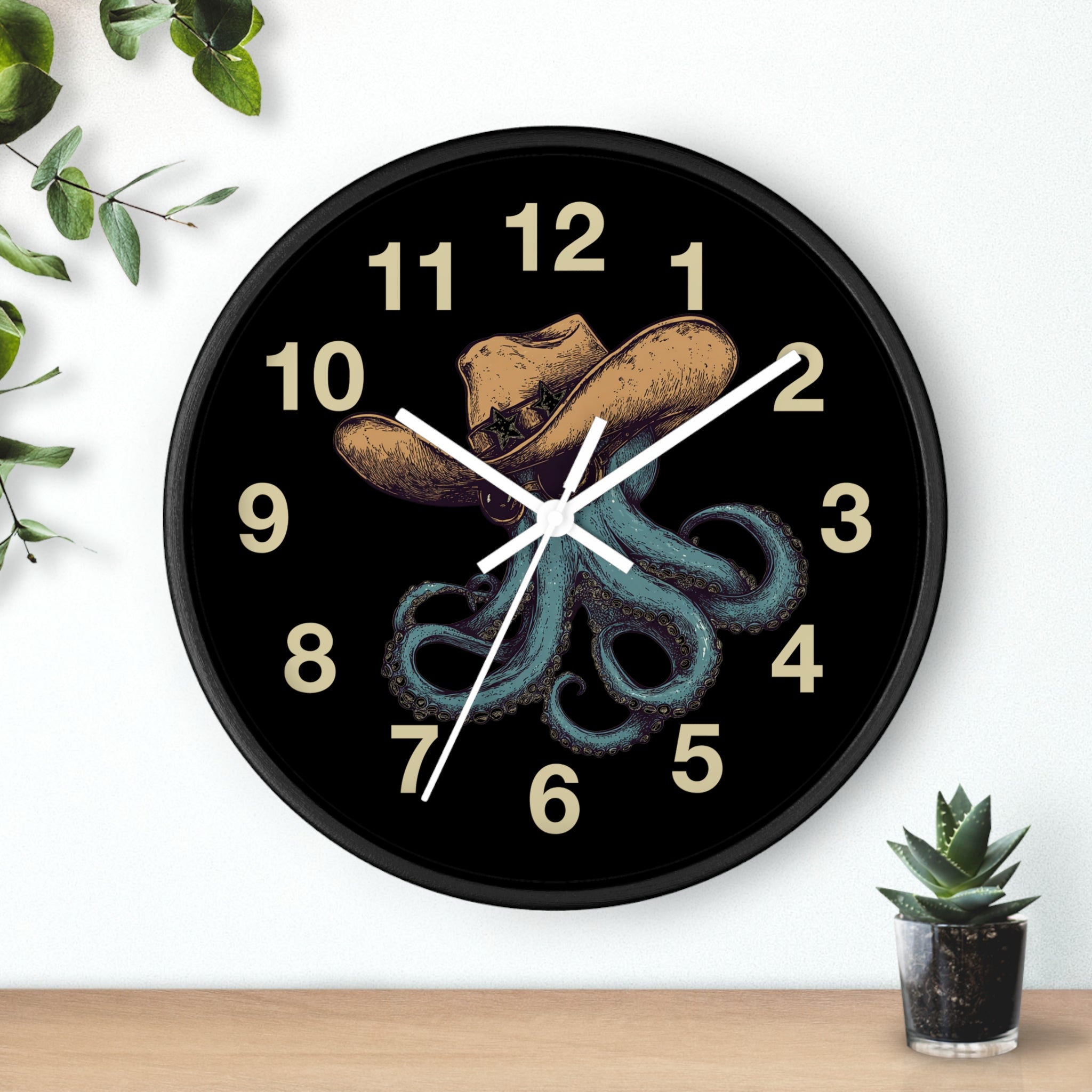 Black Wall clock with white hands and  a cowboy hat and octopus design on a black background, placed on a white wall with a plant and small pot visible.