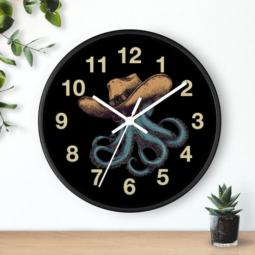 Black Wall clock with white hands and  a cowboy hat and octopus design on a black background, placed on a white wall with a plant and small pot visible.