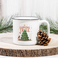 White mug with Christmas design featuring a tree and text, placed on a wooden surface with a pine cone and evergreen branches.