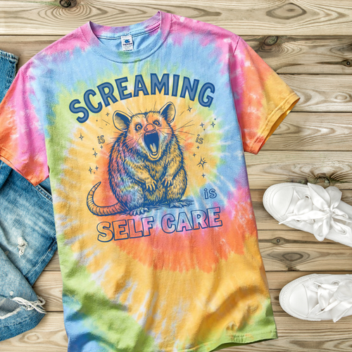Colorful tie-dye t-shirt with a graphic of an opossum and text 'Screaming is self care' on a wooden surface.