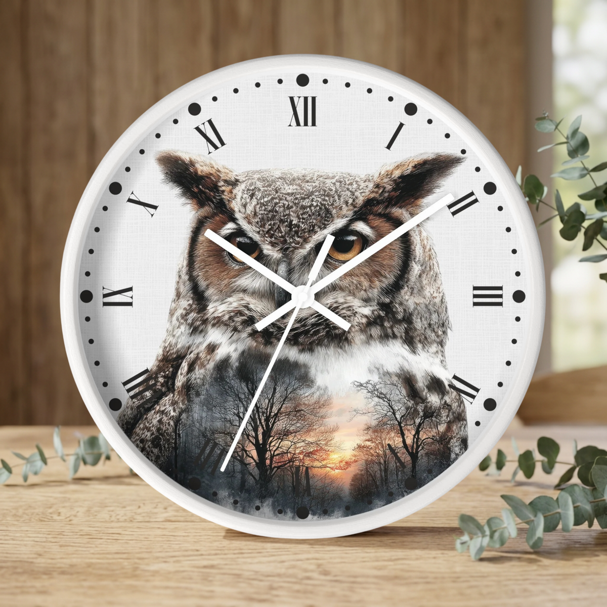 A round wall clock with a white frame and Roman numerals, featuring a large owl face over a forest sunset print on the face. The clock is shown standing on a wooden surface with decorative greenery in the background.