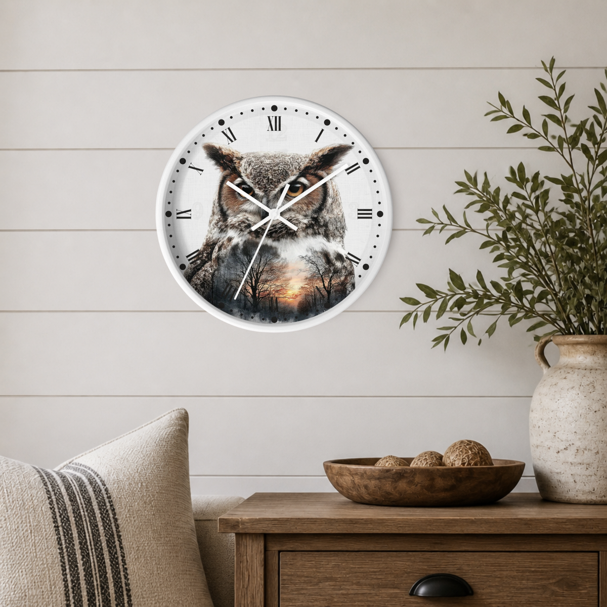 A round wall clock with a white frame and a printed owl design on the face, mounted on a light wood shiplap wall above a wooden side table with a plant and decorative bowl.