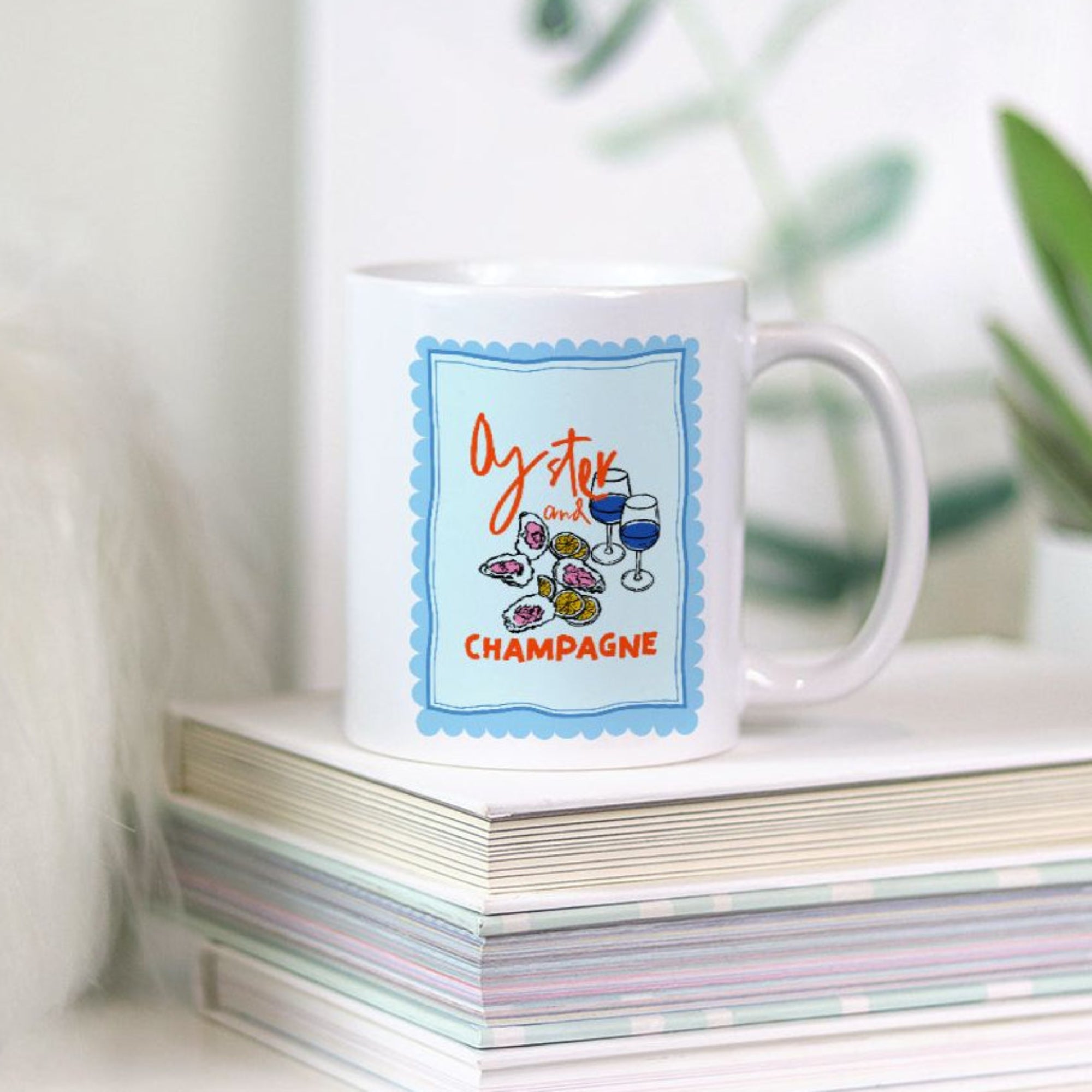 White 11oz. mug with colorful oyster and champagne design on a stack of books with a plant in the background