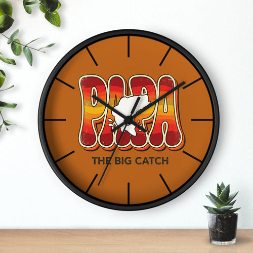 Round black wall clock with 'Papa The Big Catch' design on a white wall with a plant and small pot.