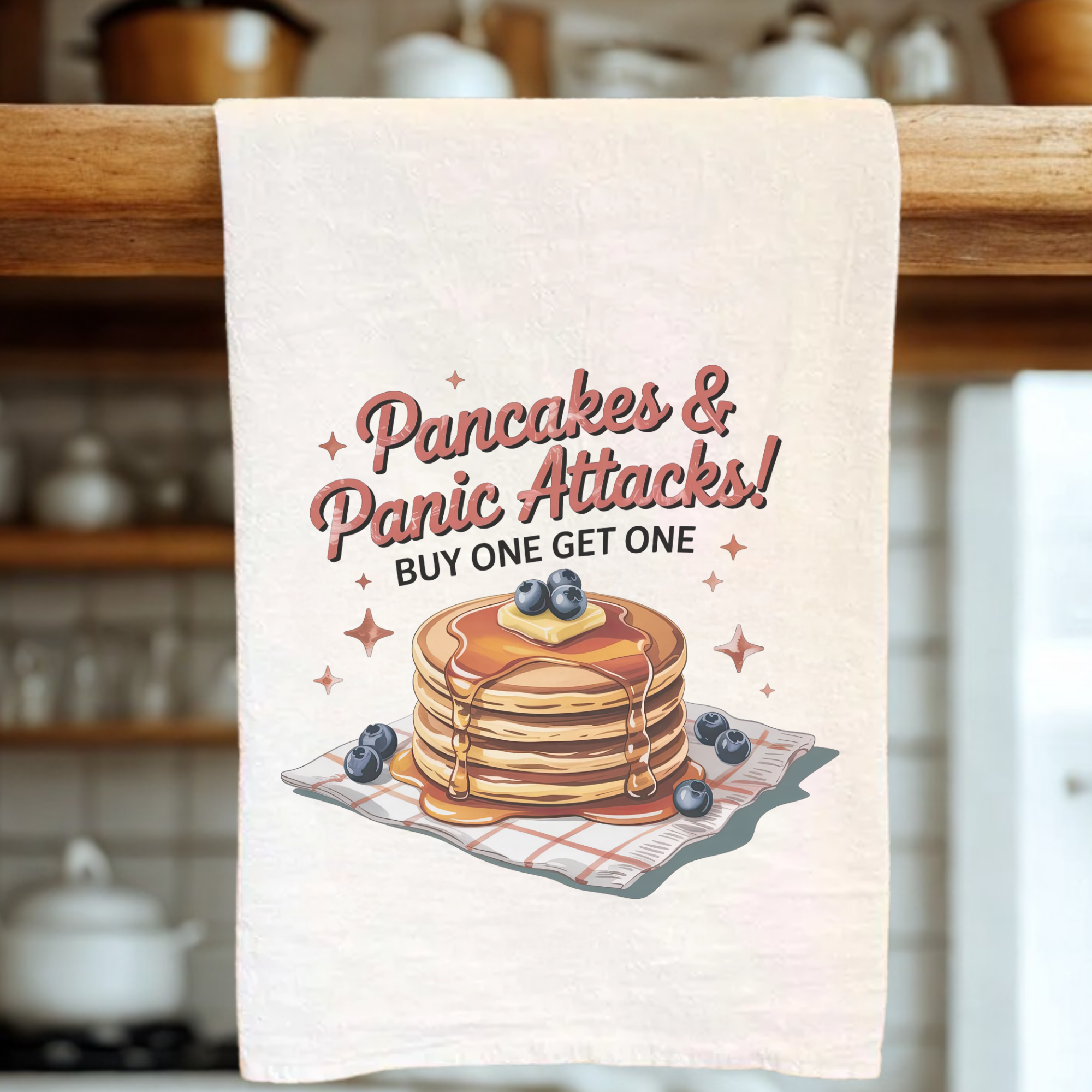 Kitchen towel with pancakes and blueberries design and text 'Pancakes & Panic Attacks! Buy One Get One' on a wooden rack.